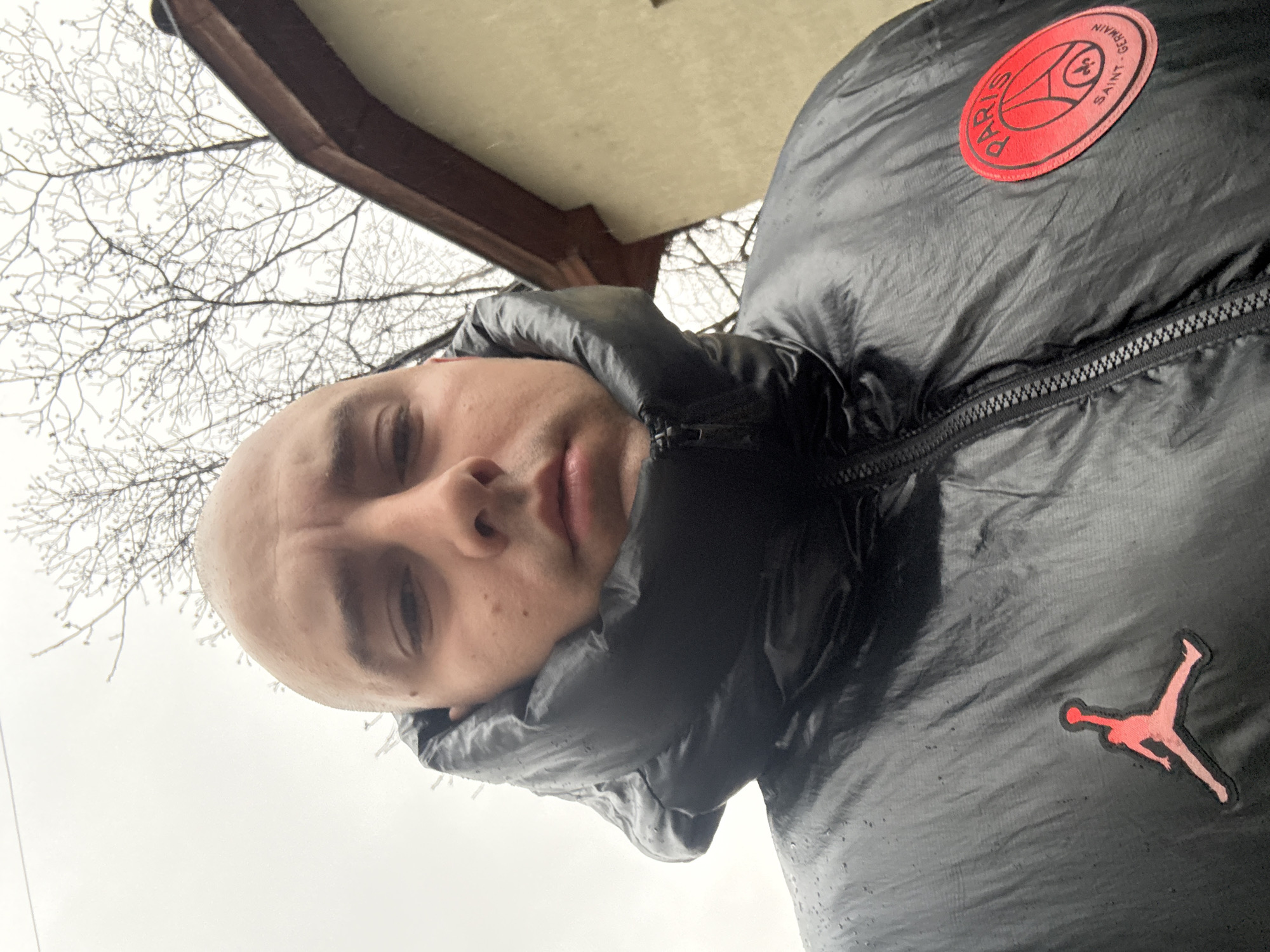 A low-angle, rotated selfie of a bald man wearing a black puffer jacket, possibly a Paris Saint-Germain (PSG) branded jacket. He is standing outdoors, potentially under a roof or awning, as indicated by a corner structure and bare branches visible against a cloudy sky. The specific location in Pleven, Bulgaria is not identifiable from the visual information.