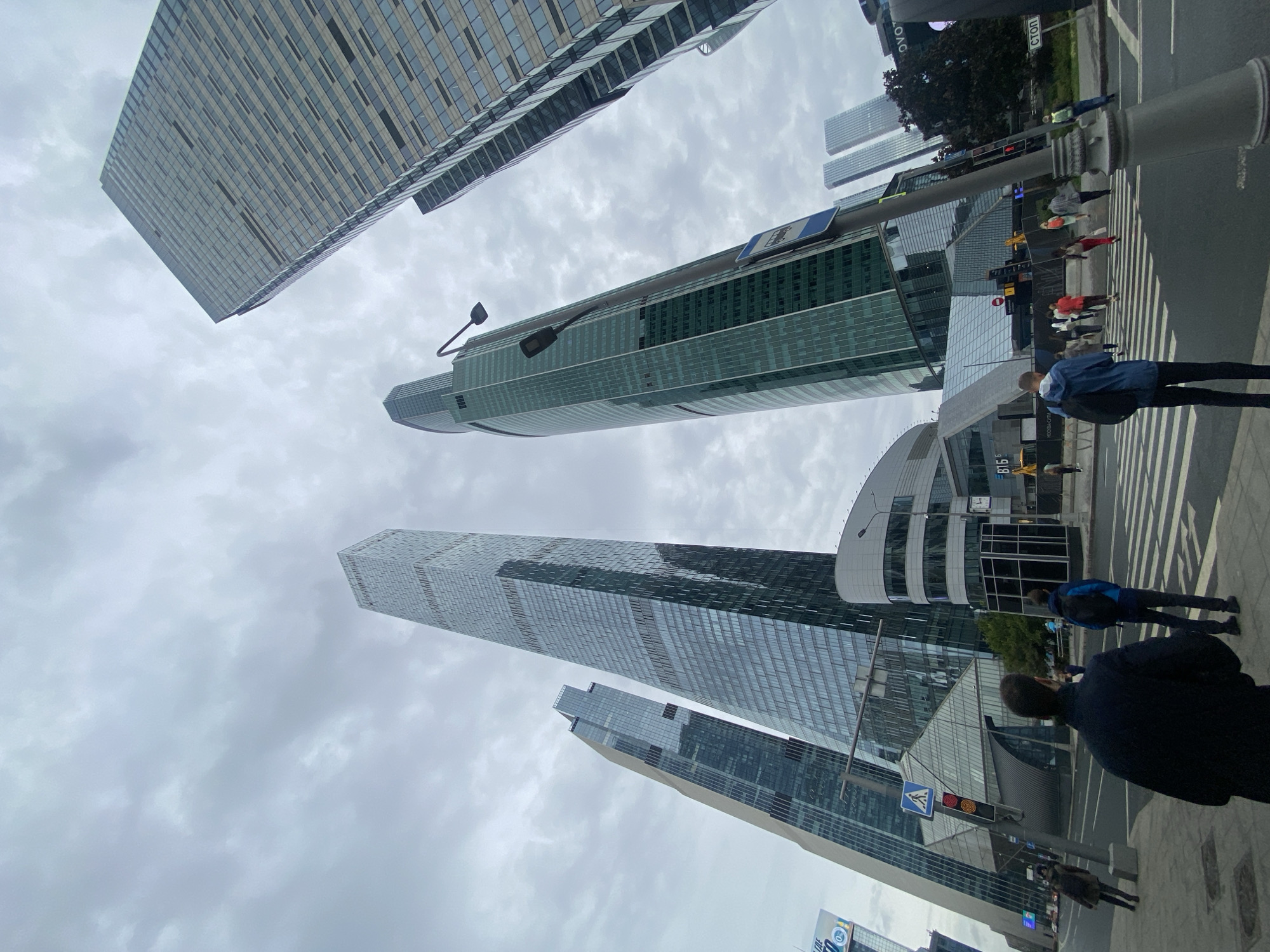 In Khimki, Russia, a wide shot captures a street-level view of modern skyscrapers under a cloudy sky. Several tall buildings with glass facades dominate the background, showcasing contemporary architectural design. In the foreground, a pedestrian crossing is visible with a few individuals walking across it and standing on the sidewalk. A traffic light with red and yellow signals is positioned on a pole, along with a bus stop sign. The scene appears to be a busy urban intersection, likely in a commercial district.