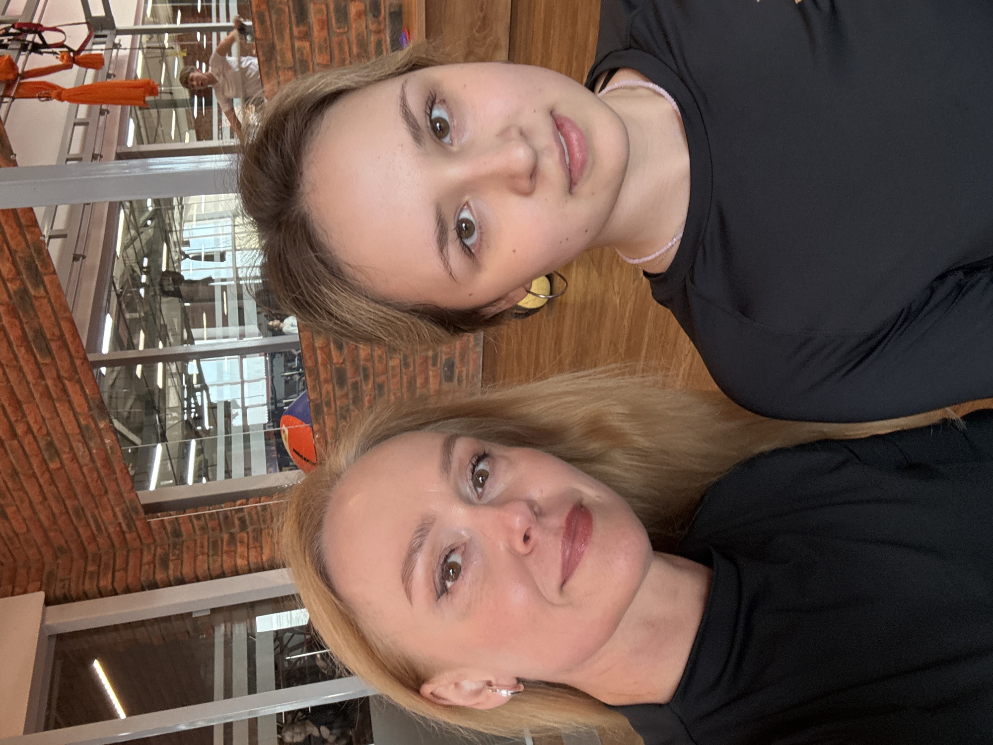 The image shows two women posing for a selfie indoors. The woman in the foreground has blonde hair, and the woman in the background has brown hair. Both are wearing black shirts.
The background reveals an interior with brick walls and windows. The architecture appears to be a multi-story building. The scene includes hints of exercise equipment or structures, possibly related to aerial yoga or a similar activity.
The location is indicated as Chaikovsky, Russia, based on the image details.