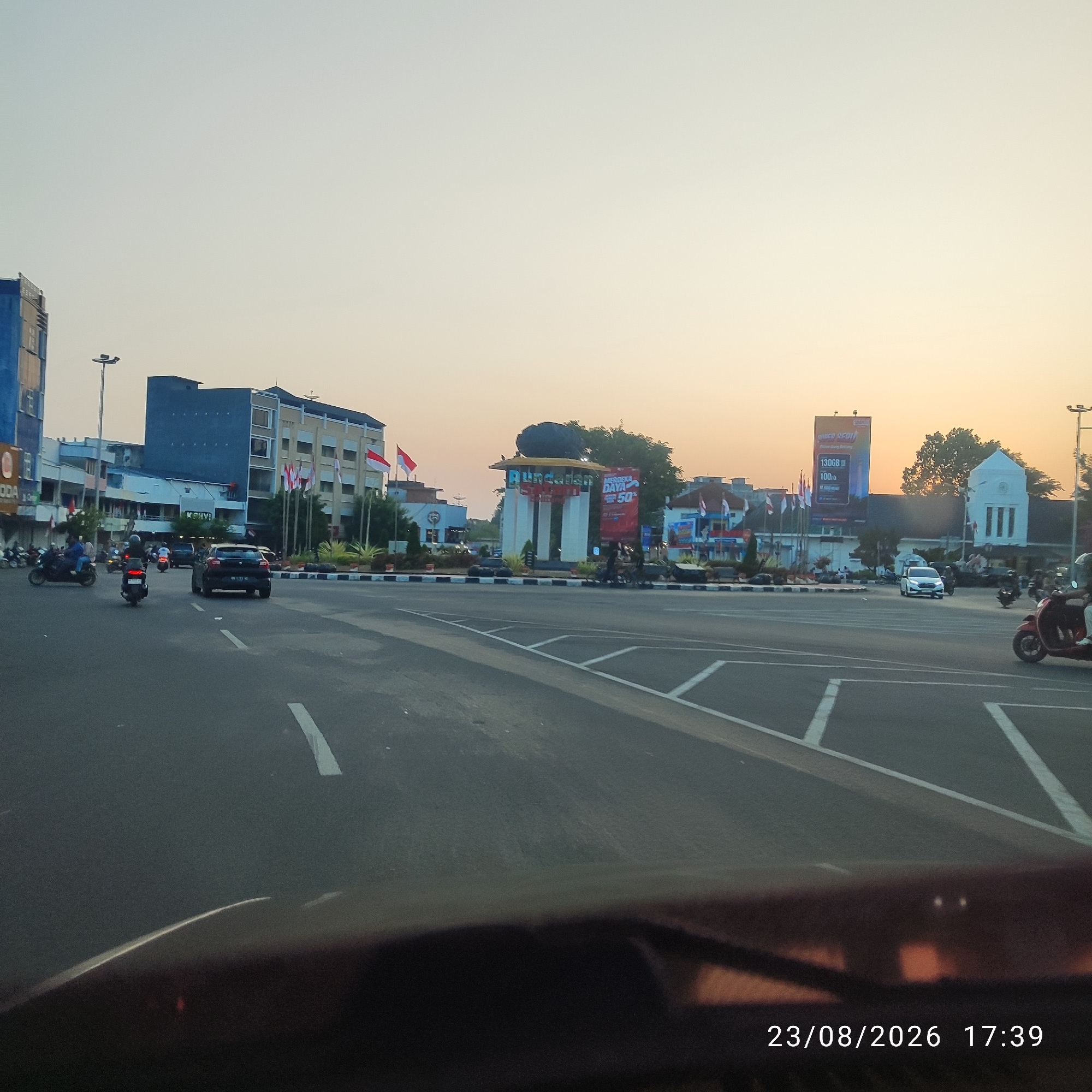The image depicts a street scene in Tanjung Pandan, Indonesia, likely at dusk due to the warm, hazy sky and the setting sun casting a soft glow. The foreground shows a road with visible lane markings and a dark, reflective surface, possibly a car's dashboard or windshield, at the bottom. In the middle ground, several vehicles, including cars and motorcycles, are in motion or parked. A prominent round-about features a monument with the inscription "Rundown," and is adorned with Indonesian flags. Buildings of various architectural styles line the street, some appearing to be commercial establishments with signage. Billboards are visible, one advertising a product with capacity specifications and another with "MERDEKA DAYA 50". The overall atmosphere suggests a typical urban environment in Indonesia during a public holiday or celebration, given the prevalence of flags. The timestamp "23/08/2026 17:39" is overlaid on the lower right portion of the image.
