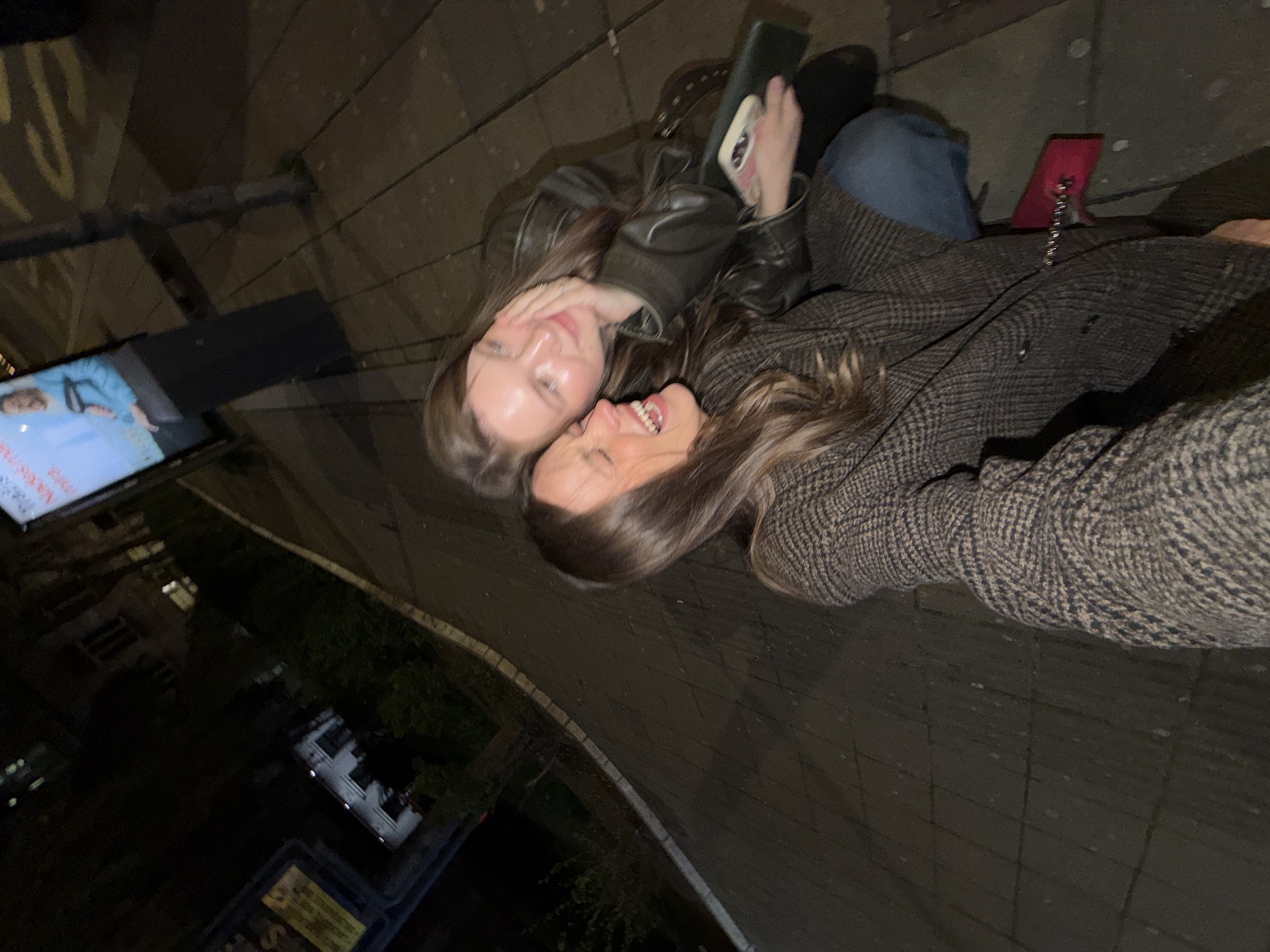 Two women are on an urban street in Belgrade, Serbia, at night. One woman is wearing a leather jacket and holding a cell phone, while the other woman is wearing a patterned coat. They appear to be interacting cheerfully and close to each other. A pink cell phone with an attached stand is visible near the woman in the patterned coat. Background elements include a large advertisement screen, a street with parked vehicles, and tree foliage.
