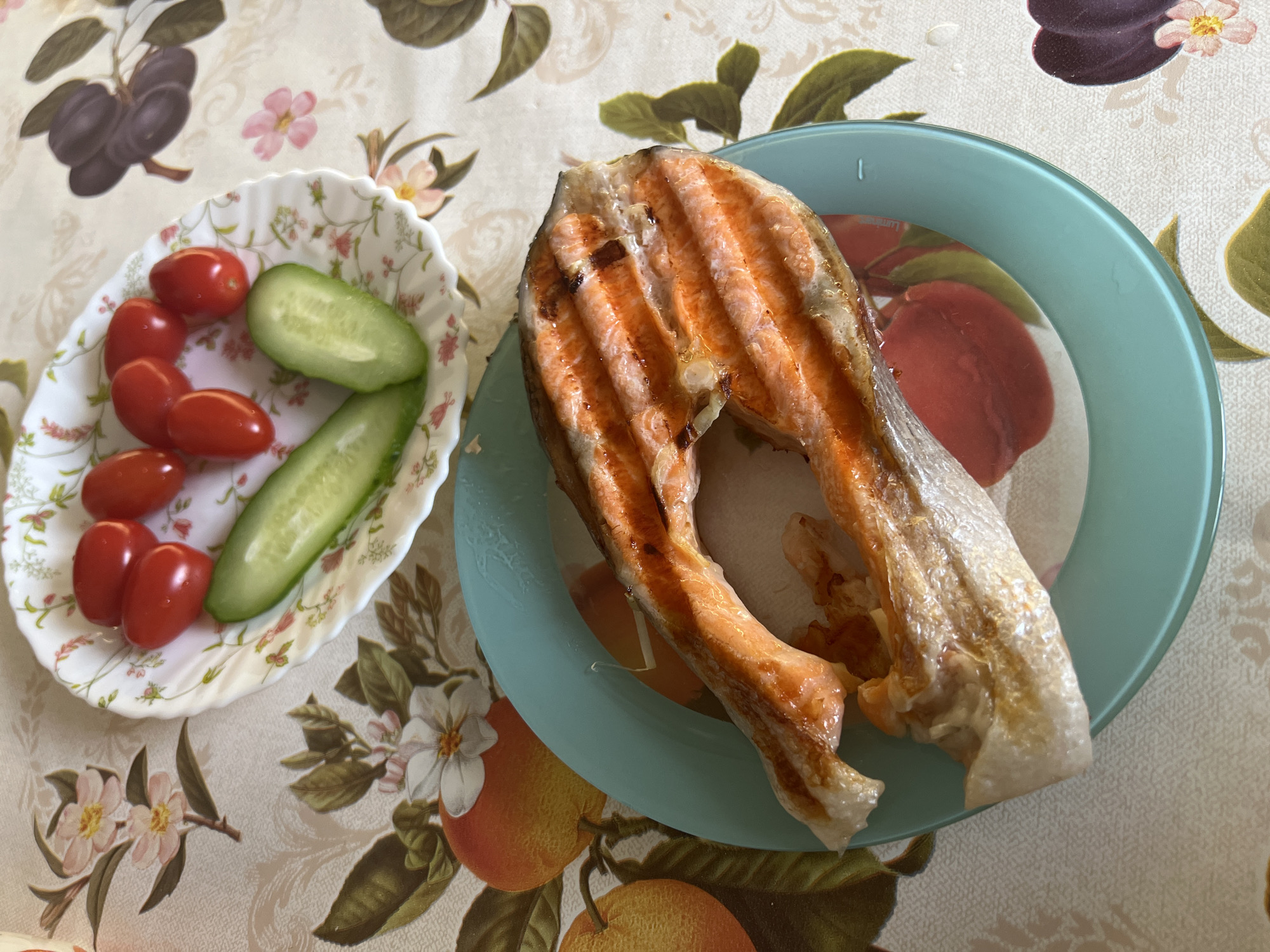 On a floral patterned tablecloth, a plate of grilled salmon lies beside a smaller plate of cherry tomatoes and cucumber slices. The scene lacks contextual clues to place it definitively in Moscow, Russia. No people are visible.