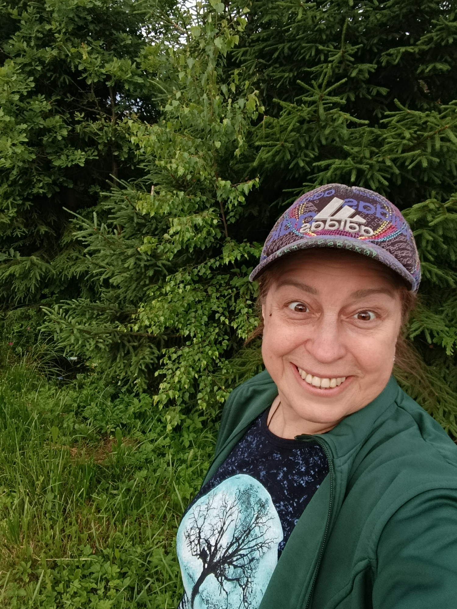 A woman smiles widely and looks directly at the camera in this close-up selfie taken outdoors. She is wearing a colorful patterned baseball cap with the Adidas logo visible. Her dark t-shirt features a graphic of a bare tree against a light blue circular background. She also wears a dark green zippered jacket. The background is filled with lush green foliage, appearing to be a dense forest or thicket, possibly in or around the city of Vereya, Russia, given the natural vegetation. The lighting suggests it is daytime.