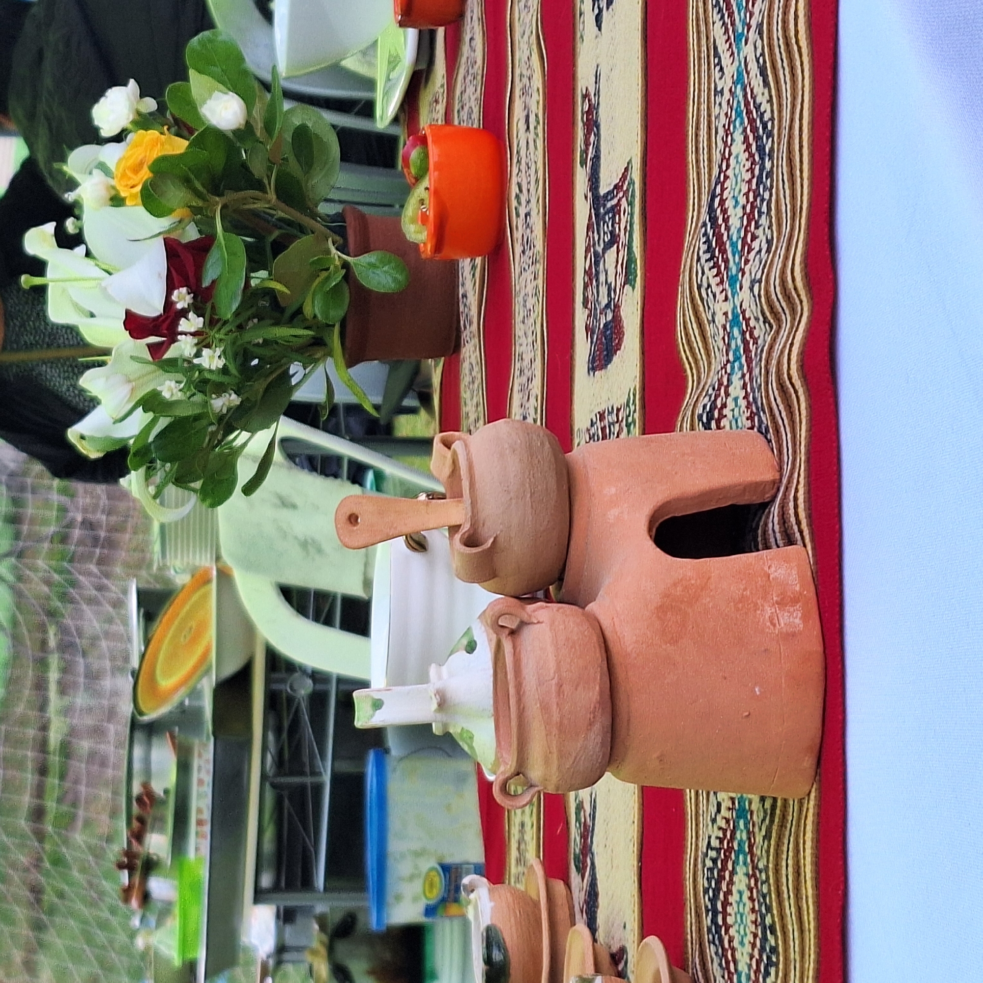The image shows a display of handmade pottery items arranged on a colorful, patterned cloth. The cloth appears to be laid out on a table or flat surface. The pottery items are primarily terracotta-colored and include vases, figurines, and other decorative pieces. A vase of flowers with white, yellow, and red blooms is visible. The background is blurred, but hints of outdoor elements suggest that the display is likely situated outside. The location is specified as Alto Selva Alegre, Peru.