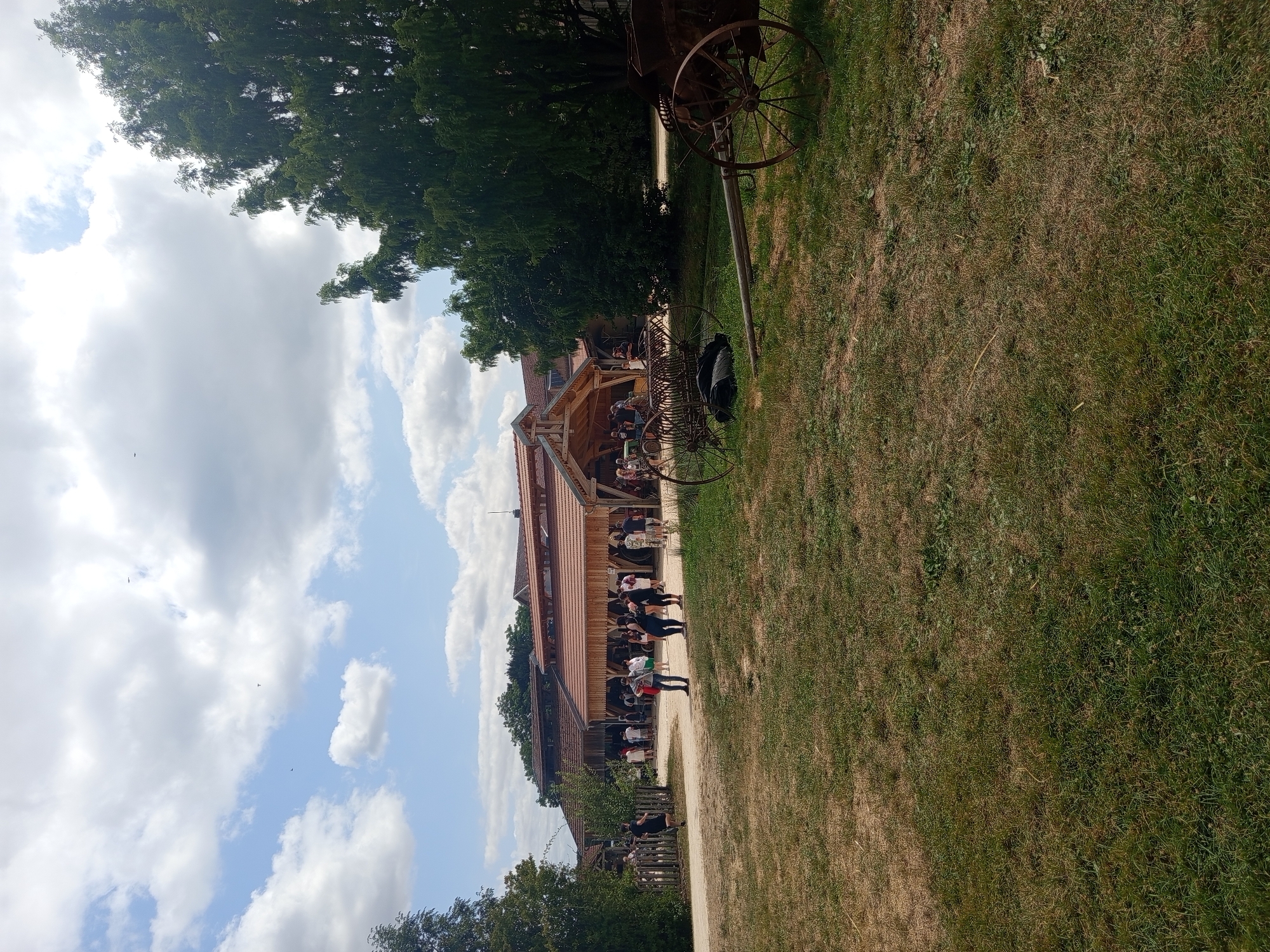 The image depicts a sunny outdoor scene at the museum of the Great War in Azannes-et-Soumazannes, France. A group of people are gathered in front of a large wooden building with a covered outdoor area. The building appears to be part of a historical exhibition or attraction. In the foreground, two pieces of antique farming equipment are present on a grassy area. The equipment includes a hay rake and a horse-drawn cart with large spoked wheels. The environment is characterized by green grass, mature trees, and a cloudy blue sky.