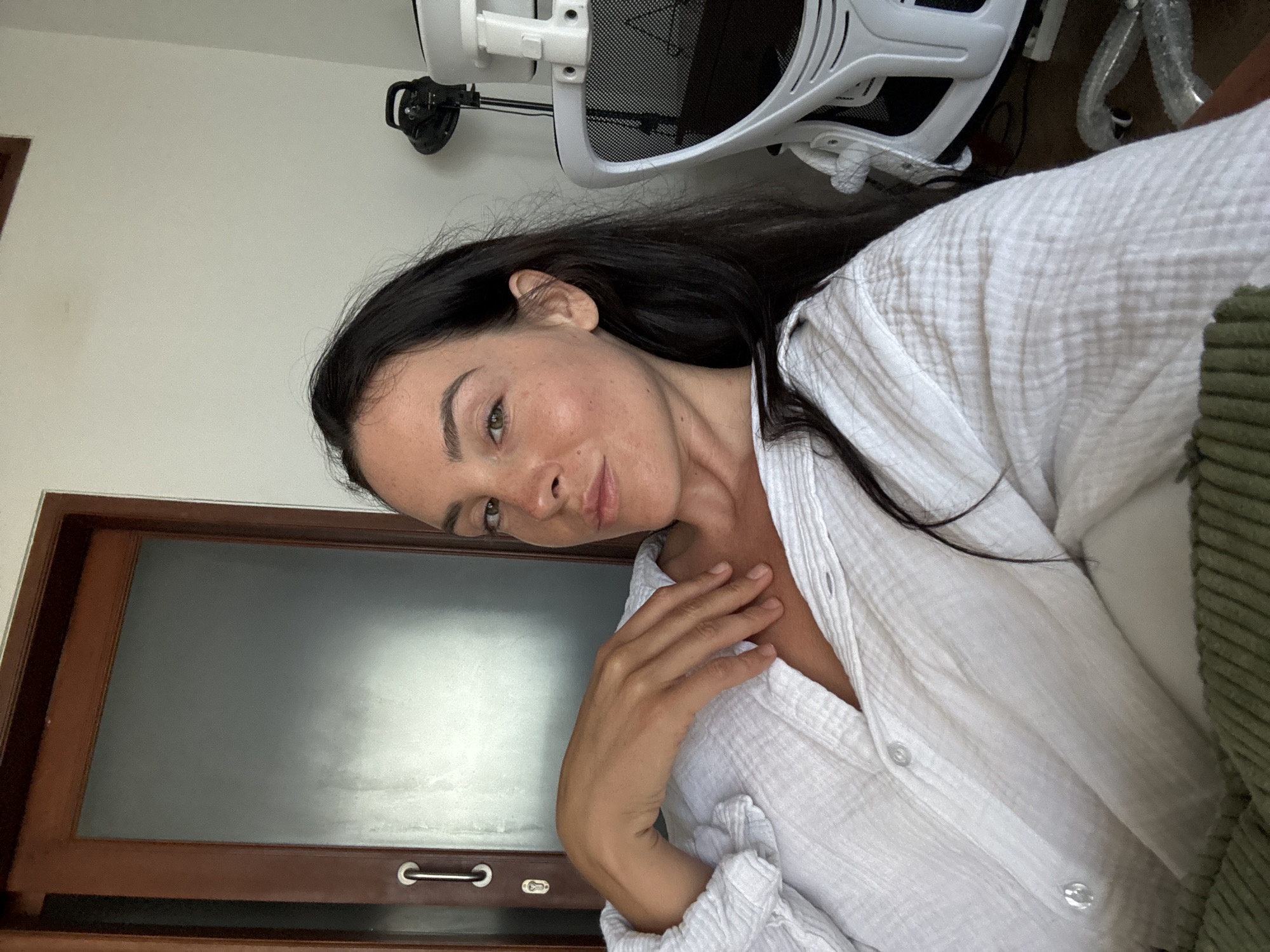 A woman with dark hair and light skin is captured in a close-up, selfie-style shot indoors. She is wearing a loose-fitting white textured shirt. Her left arm is bent, with her hand resting on her chest and collarbone. She is looking directly at the camera with a relaxed expression, her head tilted slightly. The background includes a portion of a wooden door with a frosted glass panel and a metal handle. To the right, a white office chair with a mesh back and armrests is visible, along with a small black lamp on a stand behind it. The environment suggests an indoor setting, possibly a home office or living space, located in North Kuta, Indonesia.