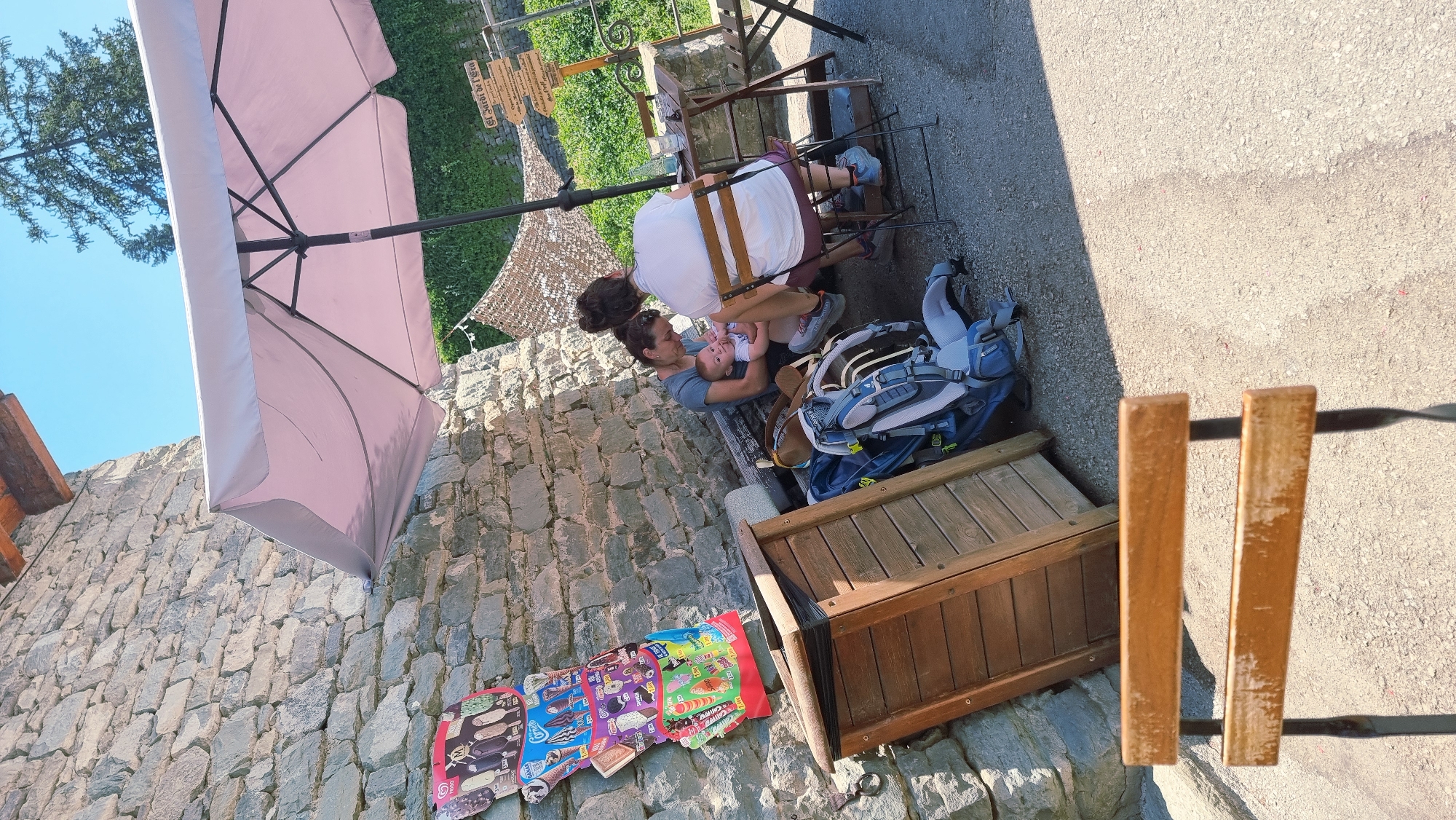 An outdoor scene captures a momentary pause for relaxation and sustenance in Rupit, Spain. In the foreground, an empty wooden chair with a metal frame stands on a gravel-covered ground. To the left, a stone wall forms the backdrop, adjacent to a wooden bin. A colorful display of ice cream advertisements is affixed to the wall above the bin. Under a light pink umbrella, two individuals and a baby are seated at a small table. One person, a woman, is holding the baby, who appears to be an infant, and is feeding it. The other individual is seated opposite them. Two backpacks, one predominantly blue and gray and the other brown, rest on the ground near the seated figures. Beyond the umbrella, a lush green hedge and a signpost are visible. The sky is a clear, bright blue.