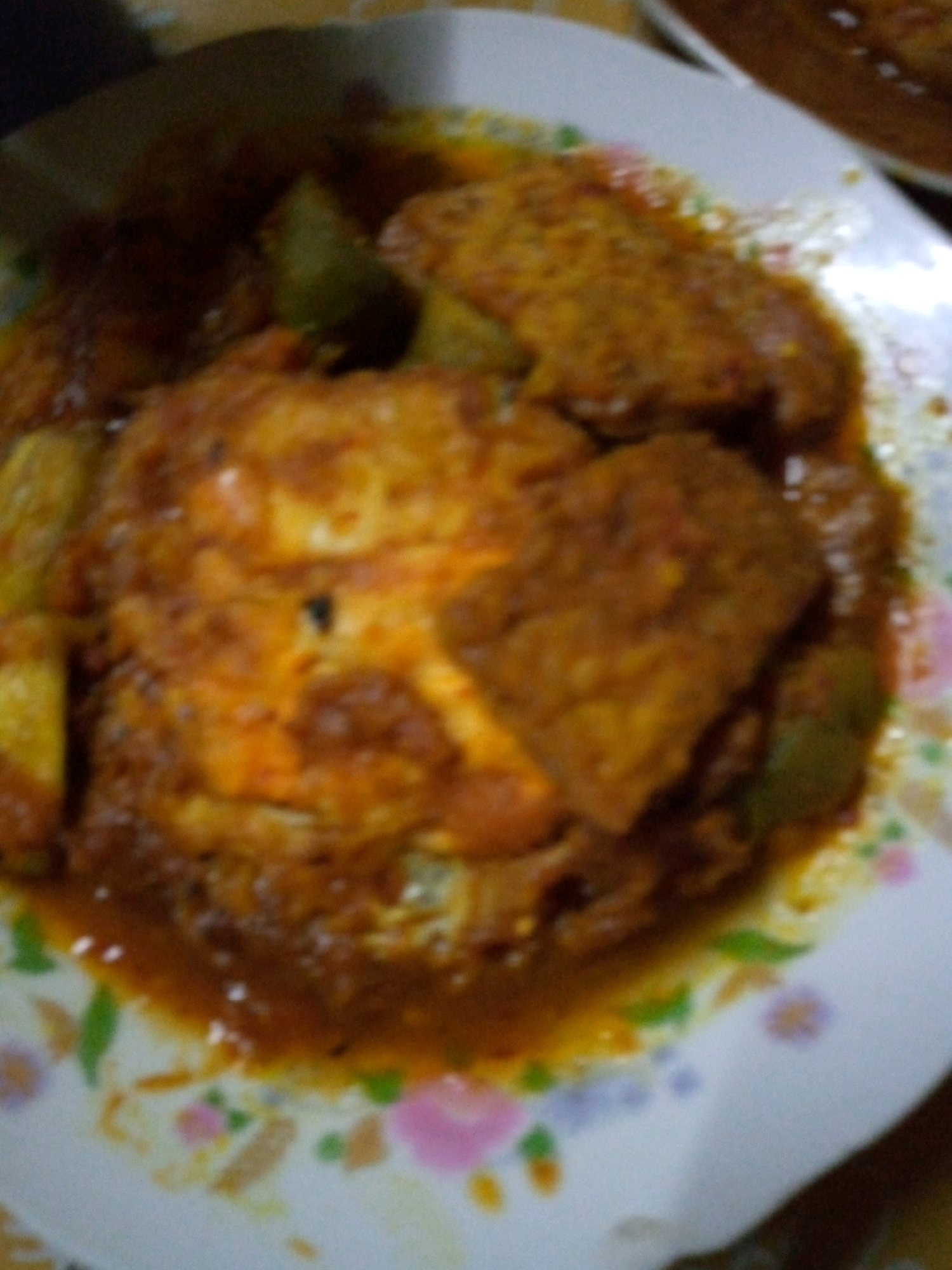 In a close-up, overhead shot, two pieces of fried fish are served in a creamy, reddish-orange sauce on a white plate with a floral pattern. The fish appears to be coated in a thick batter and is submerged in the sauce, with some green vegetables visible. The overall image is somewhat blurry, suggesting it may have been taken in low light conditions or with an older camera. The dish is presented on a table, with hints of other food or objects in the background. The scene is domestic, likely a home-cooked meal. Without further context, it is impossible to pinpoint the exact location within Sumberwono, Indonesia.