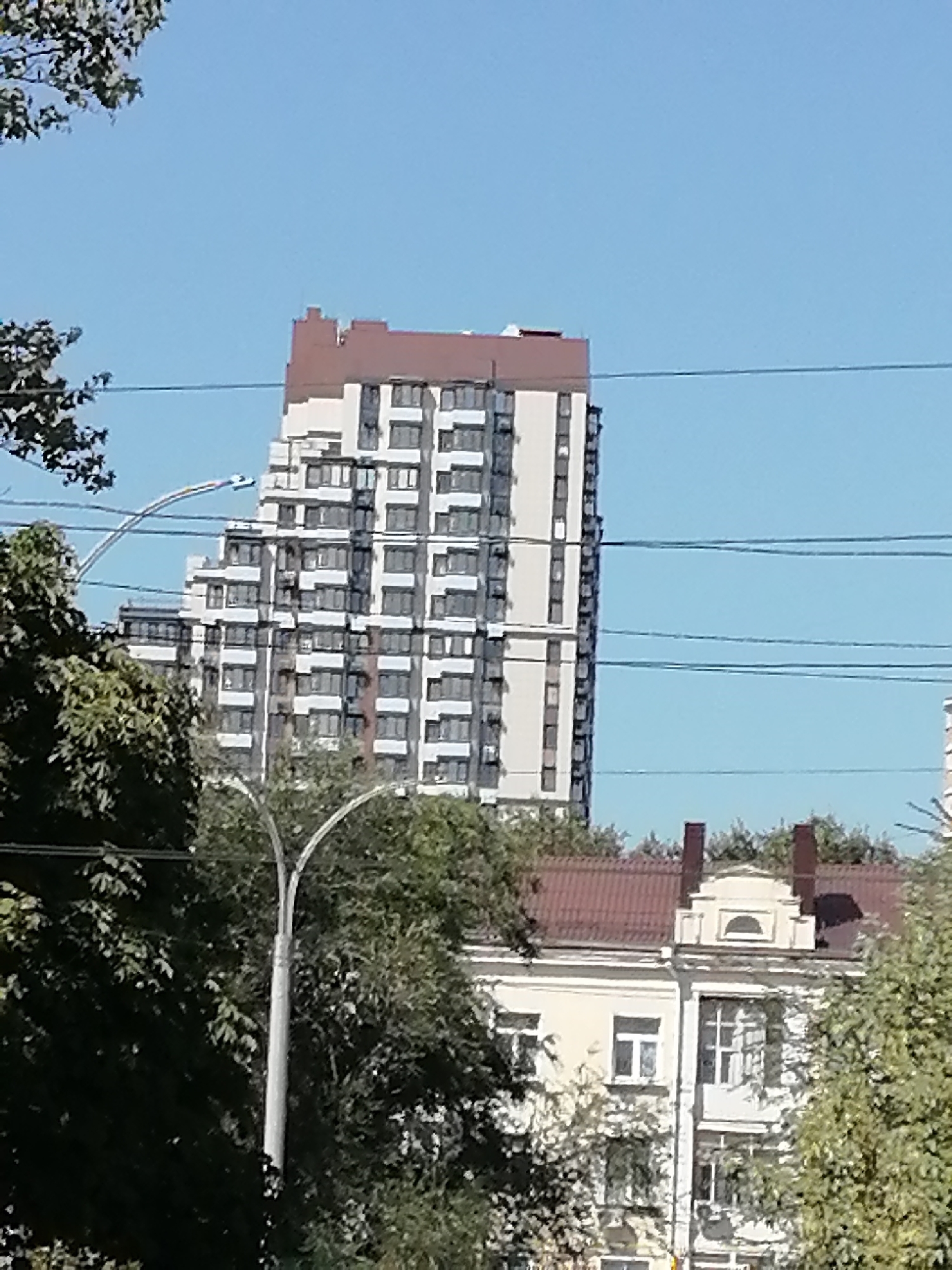 In Rostov-on-Don, Russia, a tall, modern residential building with a stepped facade and a brown roof stands against a clear blue sky. The building is partially obscured by trees and utility wires. In the foreground, a traditional, lighter-colored building with a red roof and ornate architectural details is visible, also surrounded by trees. A street lamp with a curved design is situated between the two buildings. The image captures a segment of an urban landscape, showcasing a mix of architectural styles.