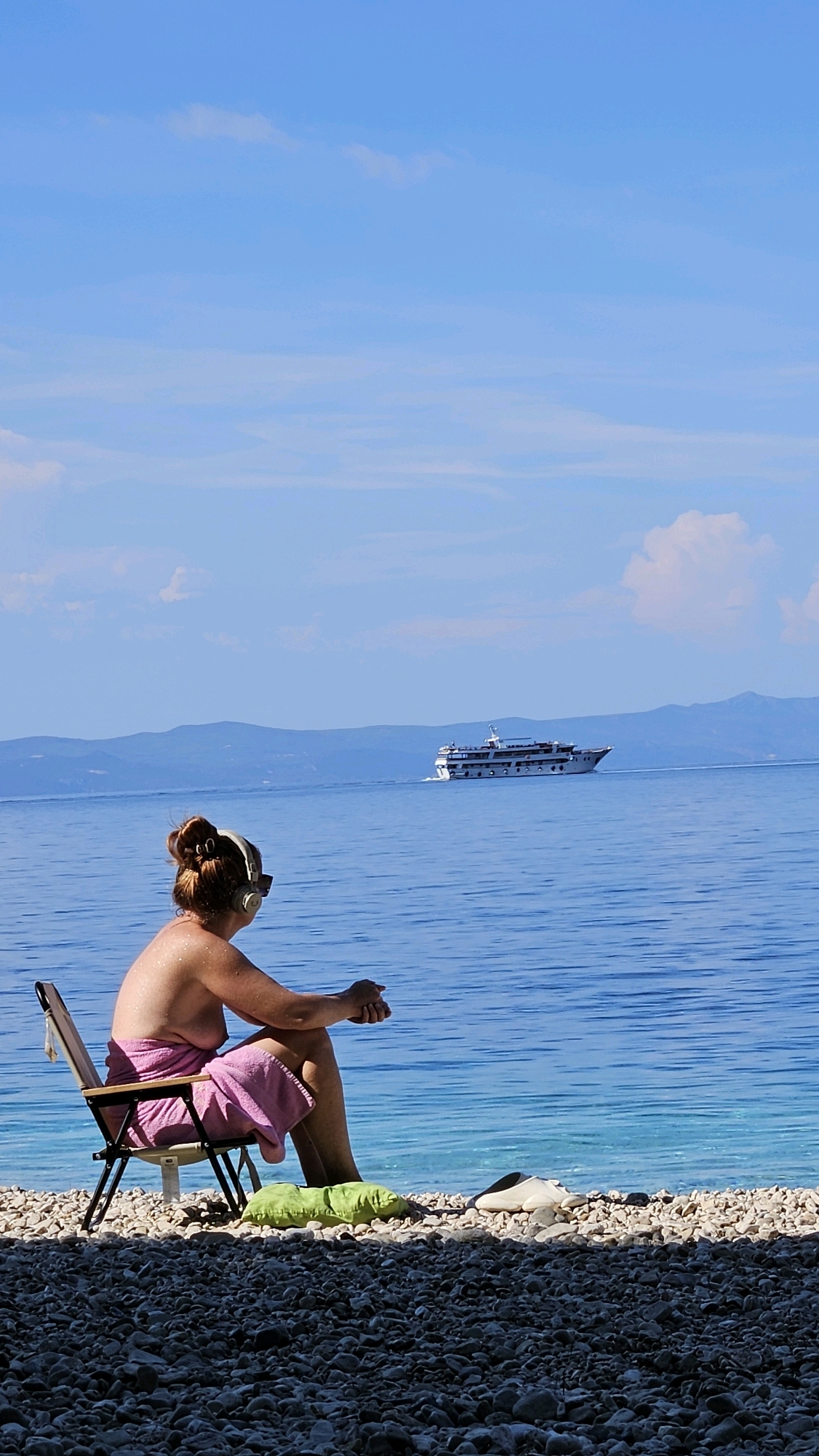 The image depicts a serene beach scene on a sunny day. In the foreground, a person sits in a foldable chair on a pebble beach, facing the blue ocean. The individual is wearing headphones and a pink towel is draped around their waist. To their right, a green pillow and a pair of white shoes are placed on the pebbles. The ocean stretches out to the horizon, with a white cruise ship visible in the distance. Mountains form a backdrop behind the ocean. The sky is a clear blue with a few wispy clouds. The location is identified as Općina Podgora, Croatia.