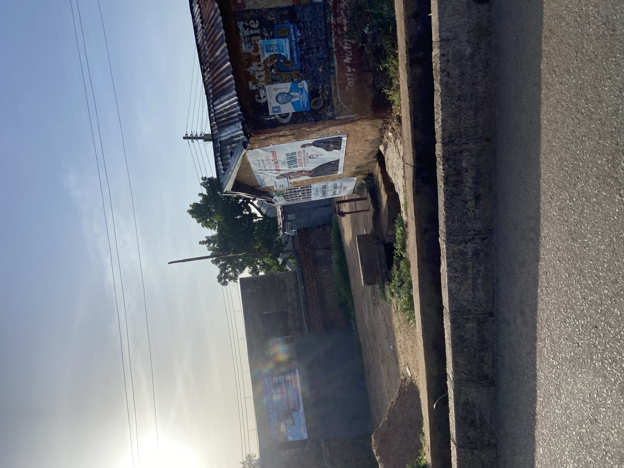 A low-angle, medium shot captures an outdoor scene on a street in Barakin Lerawi, Nigeria, under a bright, hazy sky. The foreground shows a grey asphalt road and a raised concrete curb bordering a drainage ditch. To the right of the ditch is a small, dirt area with sparse green vegetation. Beyond this, a rustic building with a corrugated metal roof is plastered with various posters and advertisements. One prominent poster features a man in a suit and the text "Together We Succeed For Joseph Nyam". Another poster displays a man's face and the words "Good Cafe". Further to the left, a dark, flat-roofed structure displays a large, colorful advertisement for "GLORIOUS LIFE INTERNATIONAL CHURCH". The overall environment suggests a developing urban or semi-urban area. No people are visible in the frame.