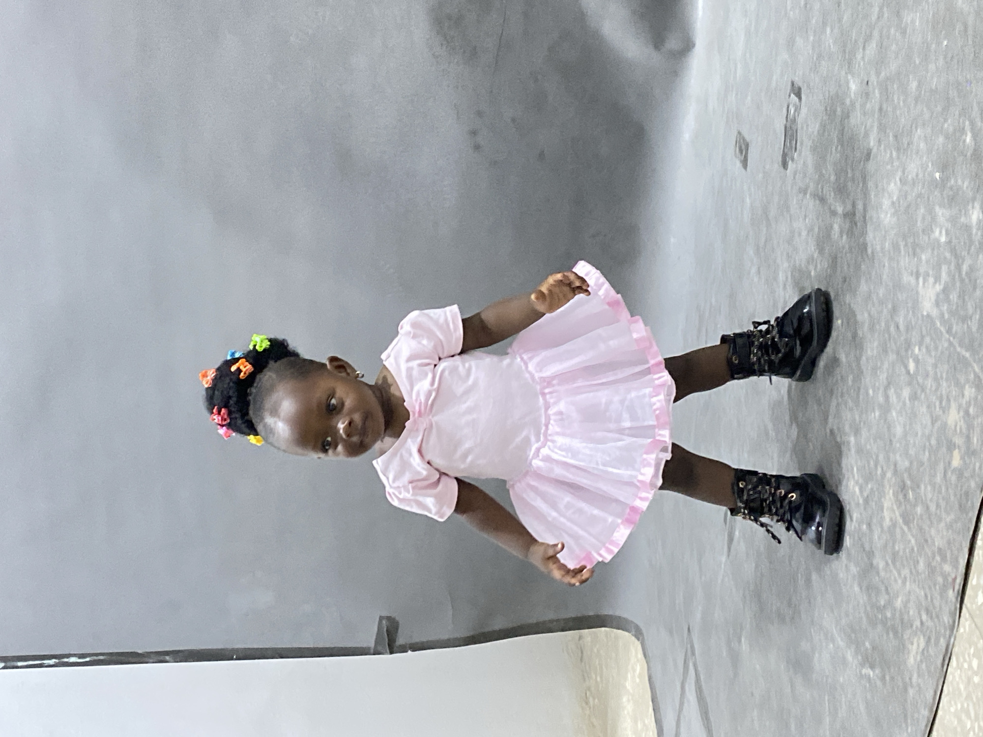 A young girl stands on a gray surface in what appears to be an indoor setting. She has dark hair styled in a high puff with colorful hair clips. She wears a light pink dress with short puffed sleeves and a frilly skirt, paired with dark, lace-up boots. The location appears to be Gura Top, Nigeria, as specified. The surface she stands on has some smudges. The background is plain, with a muted, textured gray wall.