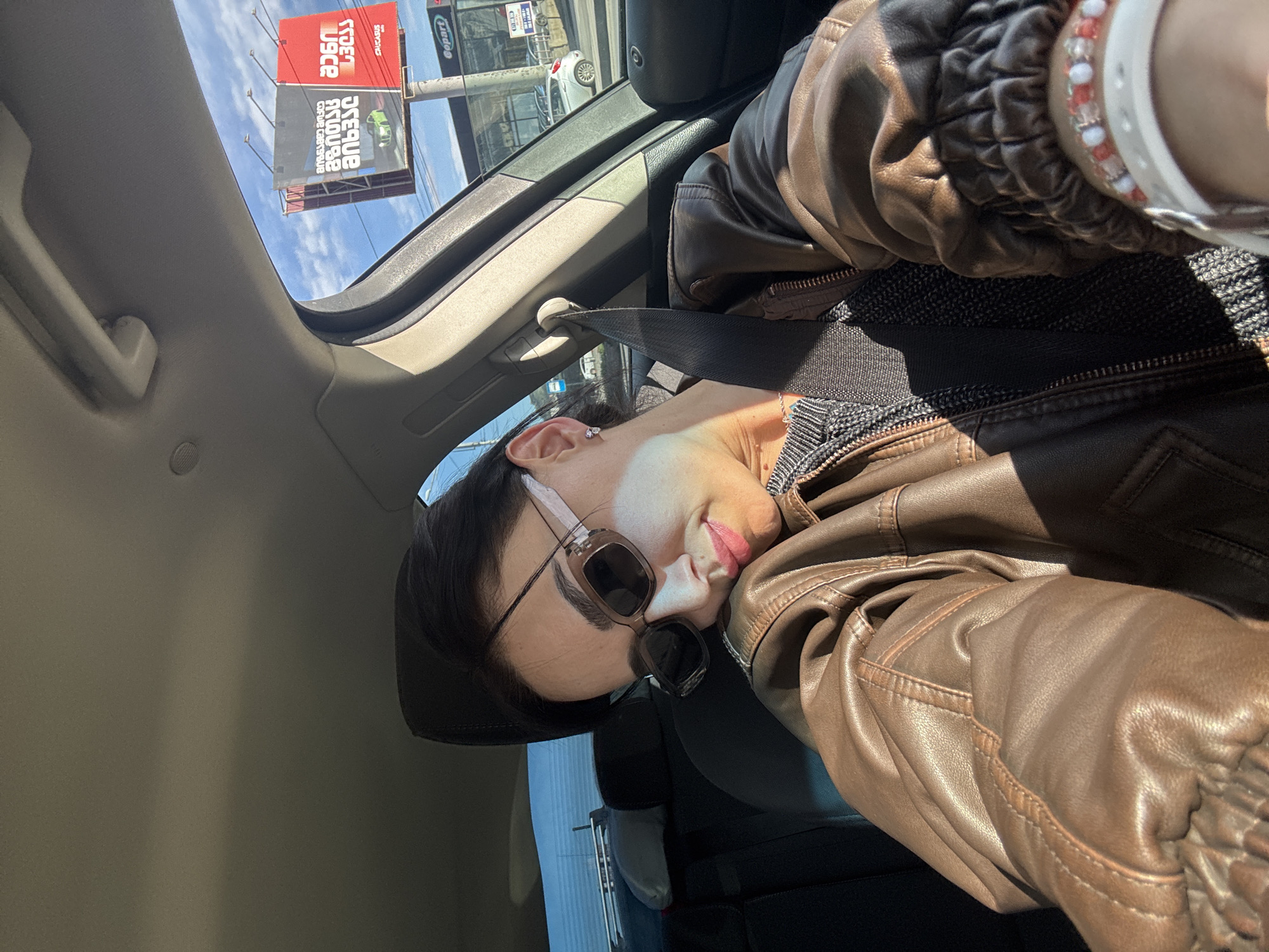 The image shows a woman inside a car, likely in Tbilisi, Georgia. She is wearing sunglasses and a brown leather jacket. A safety belt is visible. Through the sunroof, billboards with Georgian script are visible. The overall setting suggests an urban environment, potentially a commercial area, in Tbilisi.
