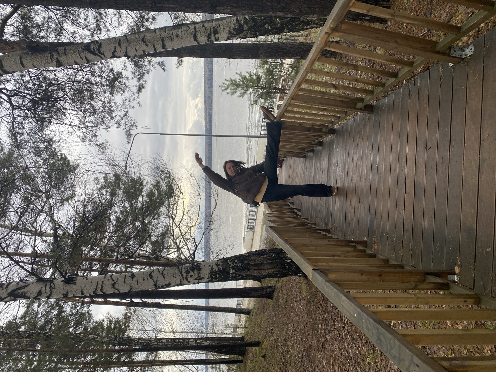 An outdoor scene captures a person performing a dynamic stretch on a wooden walkway or dock. The image, originally in portrait orientation, depicts the scene as if rotated 90 degrees clockwise. When viewed upright (landscape), a person, appearing to be a woman, is centered on a dark wooden deck, extending her right leg upwards in a high split and raising her left arm. She wears a brown jacket and dark pants.
The wooden structure features railings on the right side and appears to lead down via stairs, parts of which are visible in various crops. On the left side of the frame, a natural environment unfolds with several trees, including distinctive birch trees with white, peeling bark and some evergreen pines, framing a wide body of water in the background. The water's surface shows patches of ice or slush, indicating cold weather, possibly late autumn, winter, or early spring.
The sky above is overcast, casting a soft, diffused light over the scene. A light pole stands in the mid-ground, near the water's edge. Across the water, a distant forested shoreline is visible. The ground surrounding the wooden structure and trees is covered with fallen leaves and dry vegetation. There are some faint, illegible markings on the side support of the wooden stairs. The location is consistent with a waterfront area near Chaikovsky, Russia.