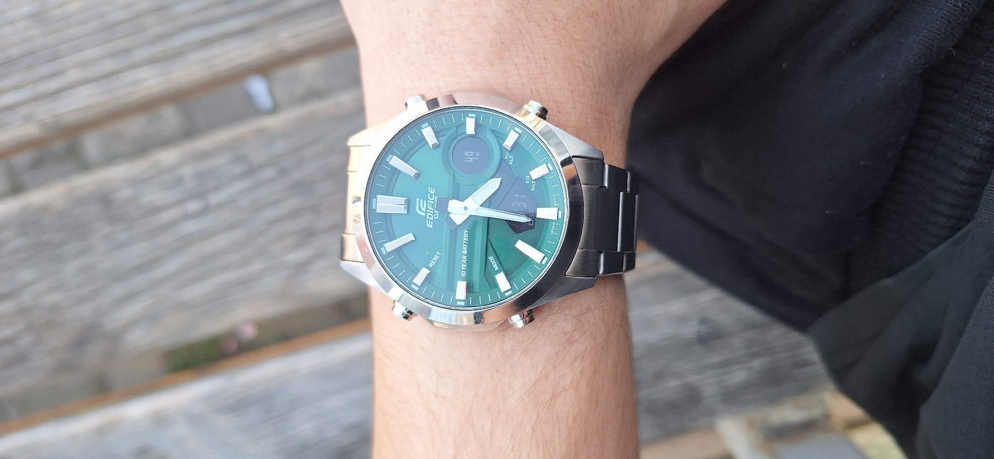 A wrist wearing a Casio Edifice watch is photographed on a wooden bench. The watch has a green face with silver and black metal casing and band. The foreground includes the wearer's arm and a dark-colored jacket. The bench is constructed of light gray wooden planks. The location is likely in Stara Zagora, Bulgaria.