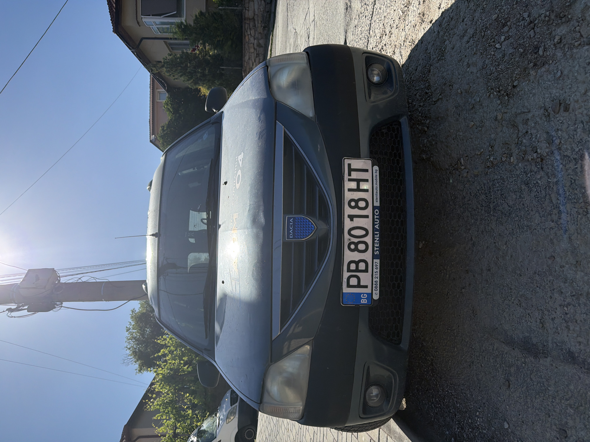 A dark gray Dacia car is parked on a gravel street in what appears to be a residential area. The car's license plate reads "PB 8018 HT" and includes the Bulgarian flag and the acronym "BG." A dealer sticker saying "STENLI AUTO" is visible on the license plate. Behind the car is a building partially obscured by trees. A power pole with numerous wires is visible to the left of the car. The sun is shining brightly, creating lens flare in the image. It is difficult to say definitively that the image was taken in Asenovgrad, Bulgaria; however, since the license plate is Bulgarian, the country is most likely Bulgaria.