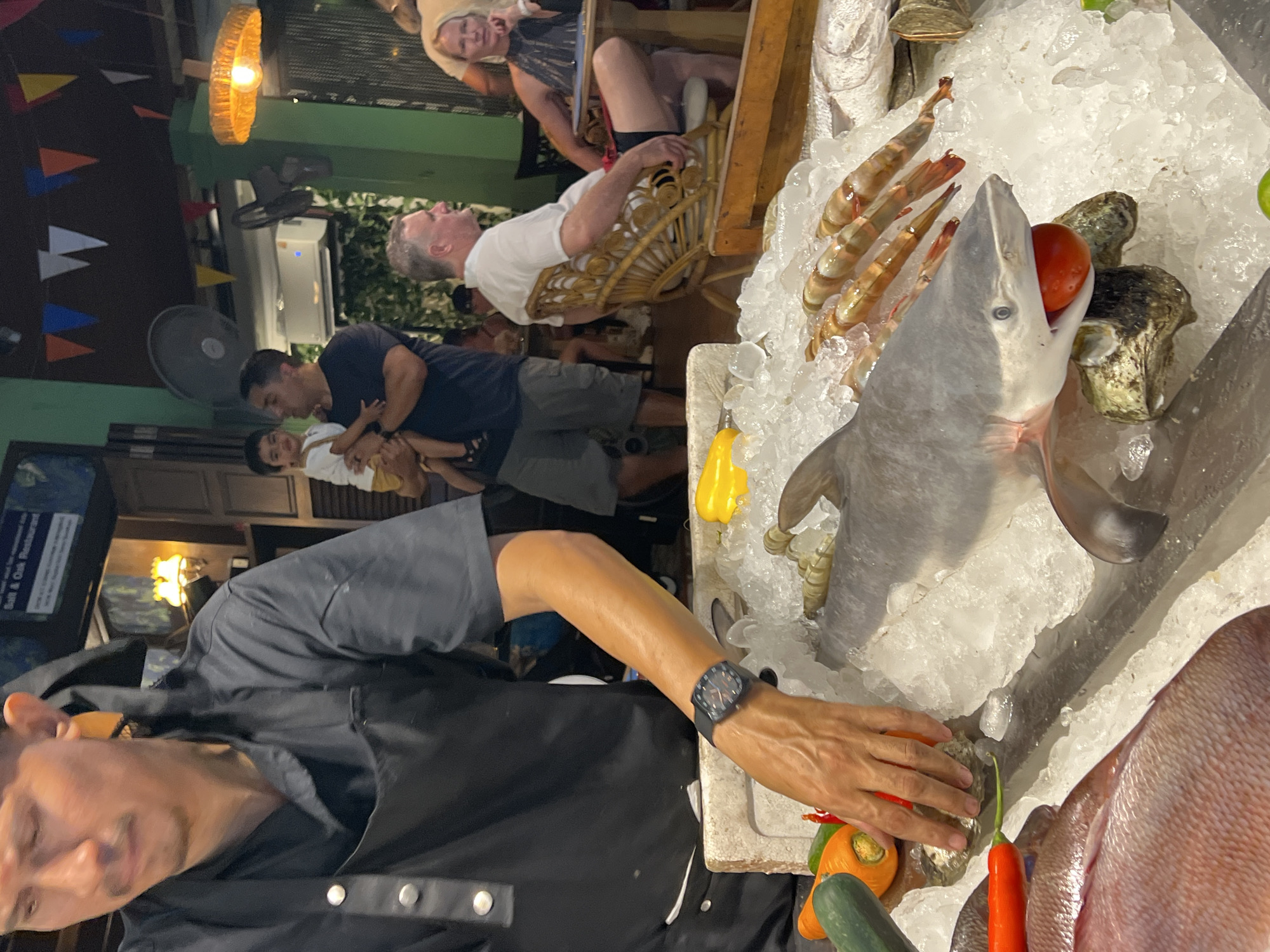In Baan Bo Phut, Thailand, a street food vendor, identifiable by his black apron and watch, arranges seafood on a bed of ice. His hand, extended over the ice, appears to be adjusting an oyster. In the foreground, a large red snapper lies on the ice, accompanied by a shark with a tomato in its mouth and several prawns. To the left, a cucumber and two orange chili peppers are visible. In the background, patrons are seated at tables, one man holding a child, while festive pennants hang overhead, suggesting a lively, open-air dining atmosphere.