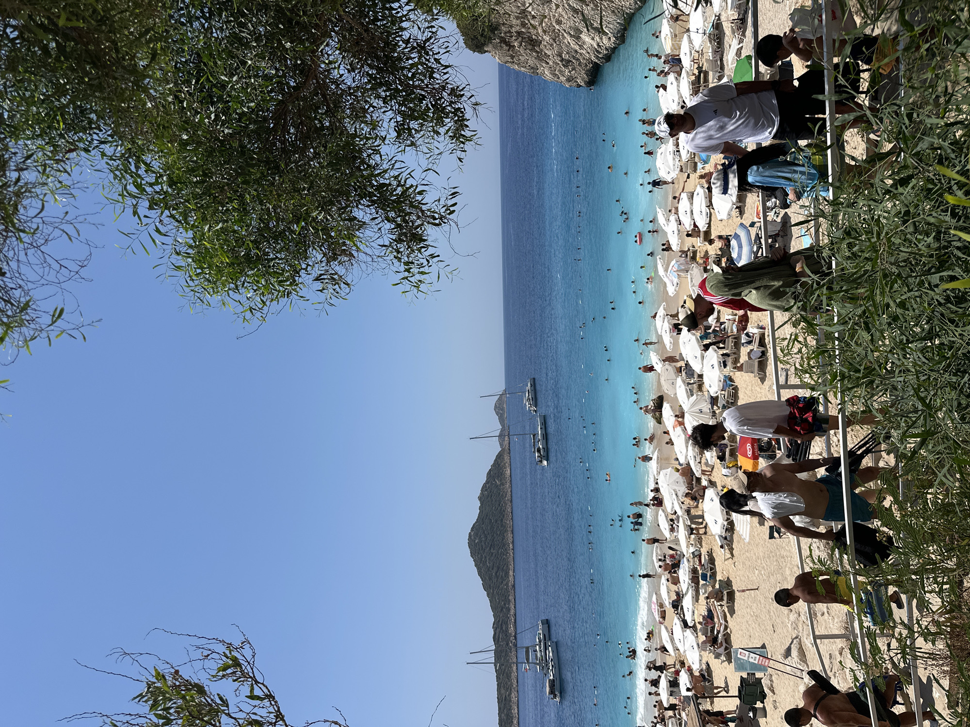 The image depicts a crowded beach scene in Kaş, Turkey, specifically Kaputaş Beach, characterized by its turquoise waters and sandy shore. The foreground shows a group of people on a set of stairs, some carrying bags and equipment, seemingly arriving at or departing from the beach. One man in the foreground is wearing a white t-shirt and a white cap, while another is wearing a green hooded towel.
The beach itself is densely populated with beachgoers relaxing under numerous white umbrellas. Many people are visible in the shallow turquoise water, swimming, floating, and enjoying the sea. Further out in the deep blue sea, several sailboats and yachts are anchored, adding to the picturesque view. A distinctive, tree-covered island or peninsula is visible in the distance, serving as a backdrop to the scenic bay. The overall atmosphere is one of a vibrant and popular summer destination. The sky is clear and blue, indicating a sunny day.