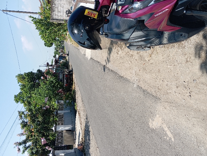 The image shows a narrow paved road with a pink Honda motorcycle parked on the right side of the road. The motorcycle is parked on the edge of the road, facing forward, with its helmet still on. The road is paved with asphalt and runs straight ahead. The road's edge is unpaved and borders a line of lush trees. There is a concrete wall and a row of buildings on the left side of the road. The houses are low-rise and have a concrete and tile exterior with an open gate leading to the yards. The sky is clear and blue, with a few clouds visible. The scene is set in a residential area in the city of Tanjunganom, Indonesia, at coordinates: -7.6, 112.035. The image was taken during the day.