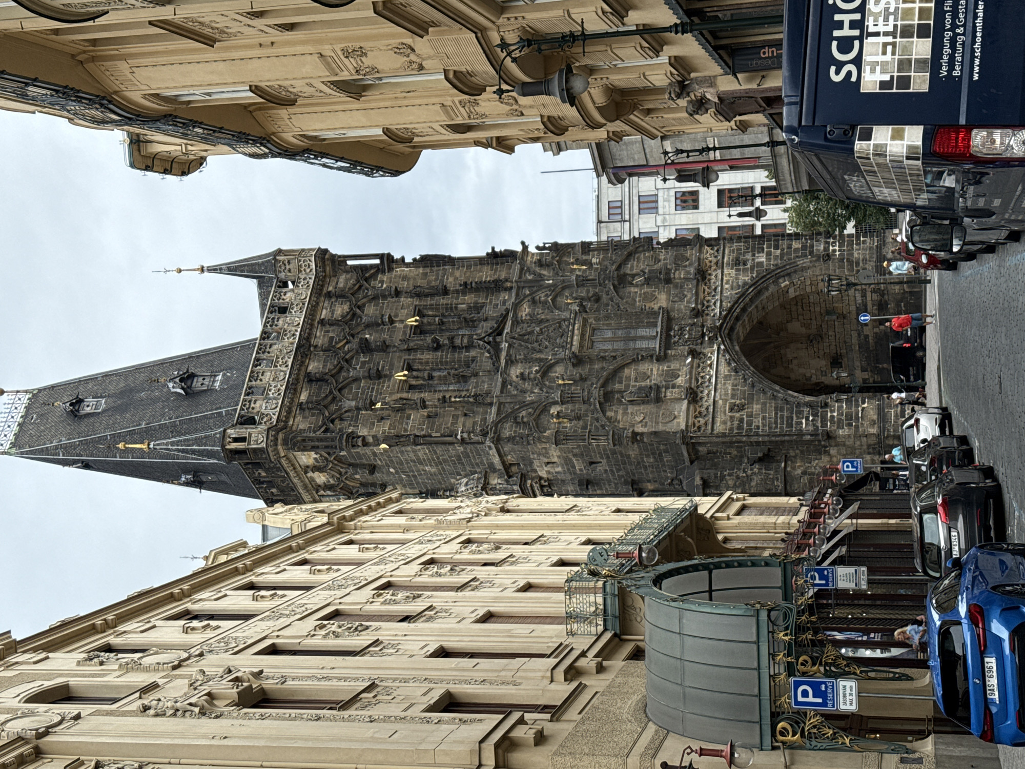 In Prague, Czechia, the image captures a street scene dominated by the Powder Tower (Prašná brána), a Gothic tower built in 1475 that served as one of the entrances to Prague's Old Town. The tower is constructed of dark stone with intricate architectural details, including statues and decorative elements. It has a tall, pointed roof topped with a spire.
Flanking the tower are two buildings. The building on the left is lighter in color and features ornate classical architectural details, including decorative relief sculptures along its facade and a green awning over an entrance. The building on the right is also a light yellow-beige color and has a curved facade with balconies and windows.
A cobblestone street runs through the foreground, lined with parked cars. A blue hatchback is parked on the left side of the street, displaying a Czech license plate. To its right are several other cars, including a dark gray hatchback and a white sedan. A dark blue van with white lettering and a mosaic-like pattern is parked further down the street on the right.
Several people are visible in the scene. A woman in a light blue top stands near the entrance of the building on the left. A man in a red shirt and blue shorts stands on the sidewalk in front of the archway of the Powder Tower. Other individuals are walking or standing near the cars and buildings.
The sky is overcast, suggesting a cloudy day. Streetlights and signage are visible along the street, including "P" parking signs. The overall impression is a bustling urban environment with historical architecture.