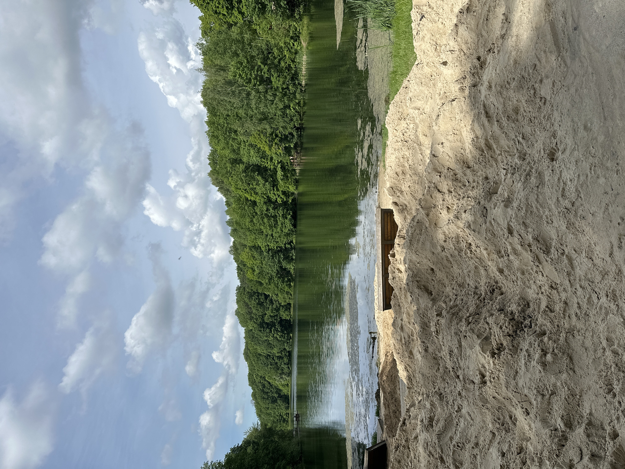 A landscape photograph depicts a serene lake scene. The image is divided vertically: on the left, a cloudy sky hangs over a line of dense green trees that stretch along the lake's edge. The water is calm with reflections of the trees. On the right, a large pile of sand dominates, covering the bottom half of the image. The image is taken near a sand pit area along the river bank in the city of Nizhny Novgorod, Russia.