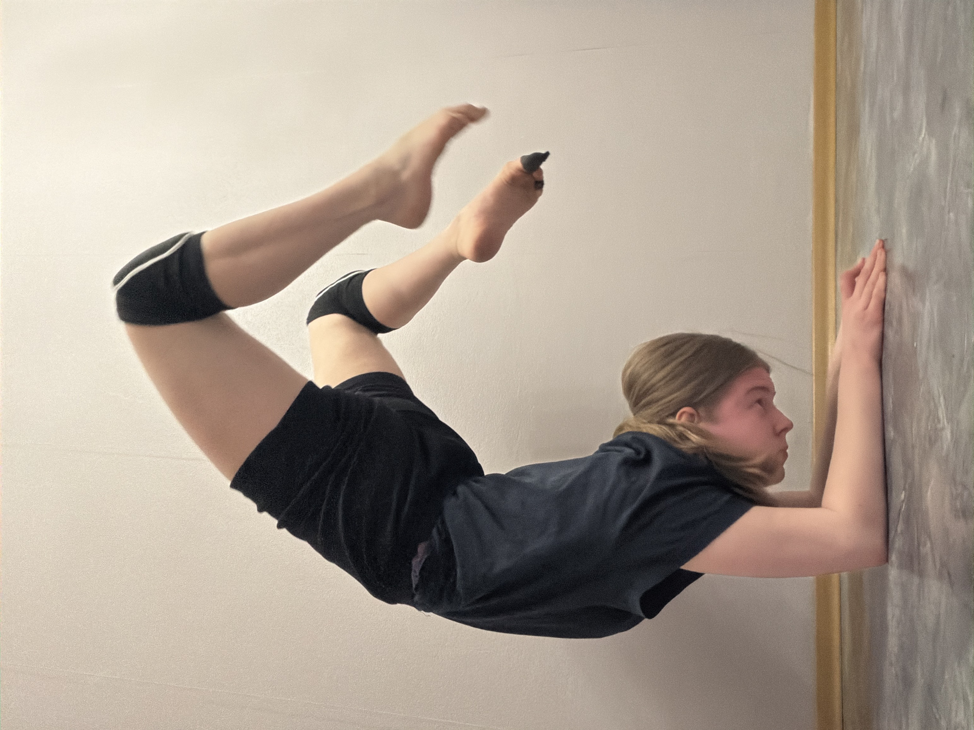 A young person, likely female, is performing a wall-assisted handstand or a similar yoga pose against a wall. The person is wearing black athletic shorts with white trim, black knee pads, and a dark-colored t-shirt. They are barefoot, with a dark covering on one toe. The wall is pale with a dark, textured panel. The location is not discernible from the image.