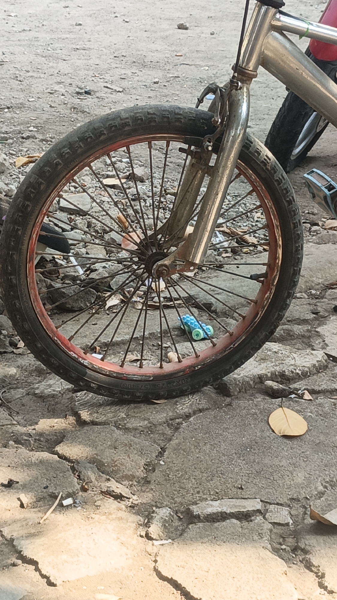 The image shows a close-up of a bicycle wheel on a cracked concrete surface. The wheel has a red rim and many spokes. A portion of the bicycle frame, including the handlebars and front fork, is visible. The background consists of a rough, gravelly surface. There is a small, blue and green toy car on the ground near the wheel. Scattered debris, including leaves and small stones, is present on the ground. The scene appears to be outdoors in daylight.