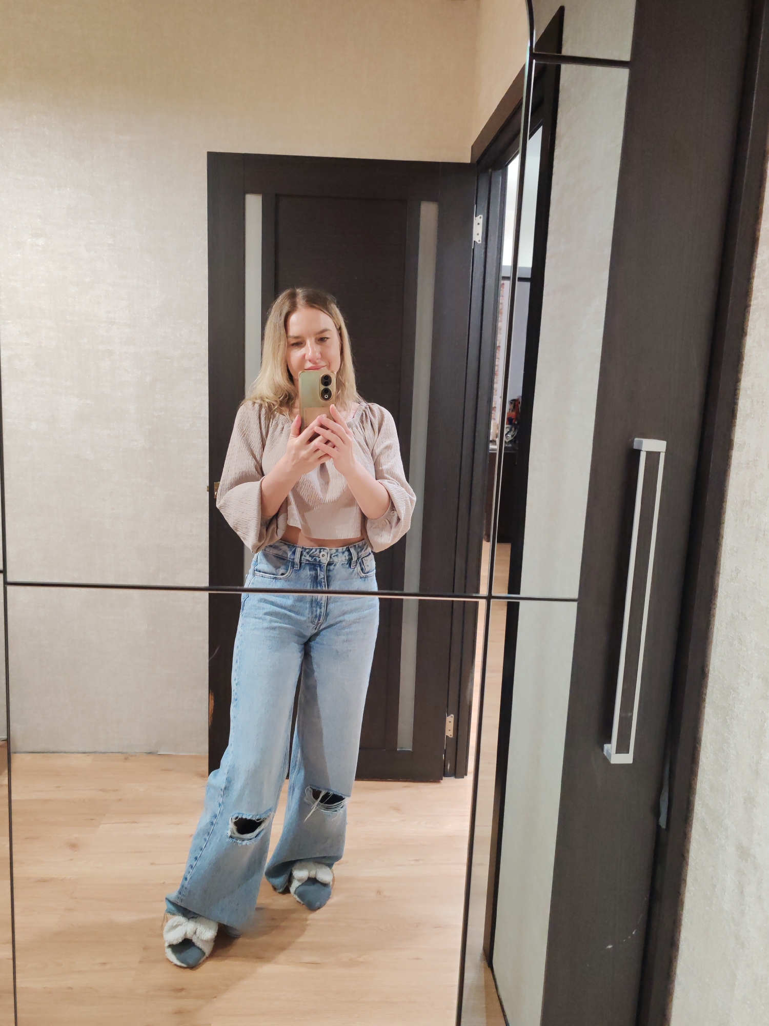 A woman is taking a selfie in a mirror. She is wearing a beige top, ripped light blue jeans, and light blue slippers with bows. The background shows a room with light walls and a dark brown door. The room appears to be indoors, possibly in a house or apartment. The location is not discernible from the image. It is not possible to identify if the location is within the city of Petropavlovsk-Kamchatsky, Russia.