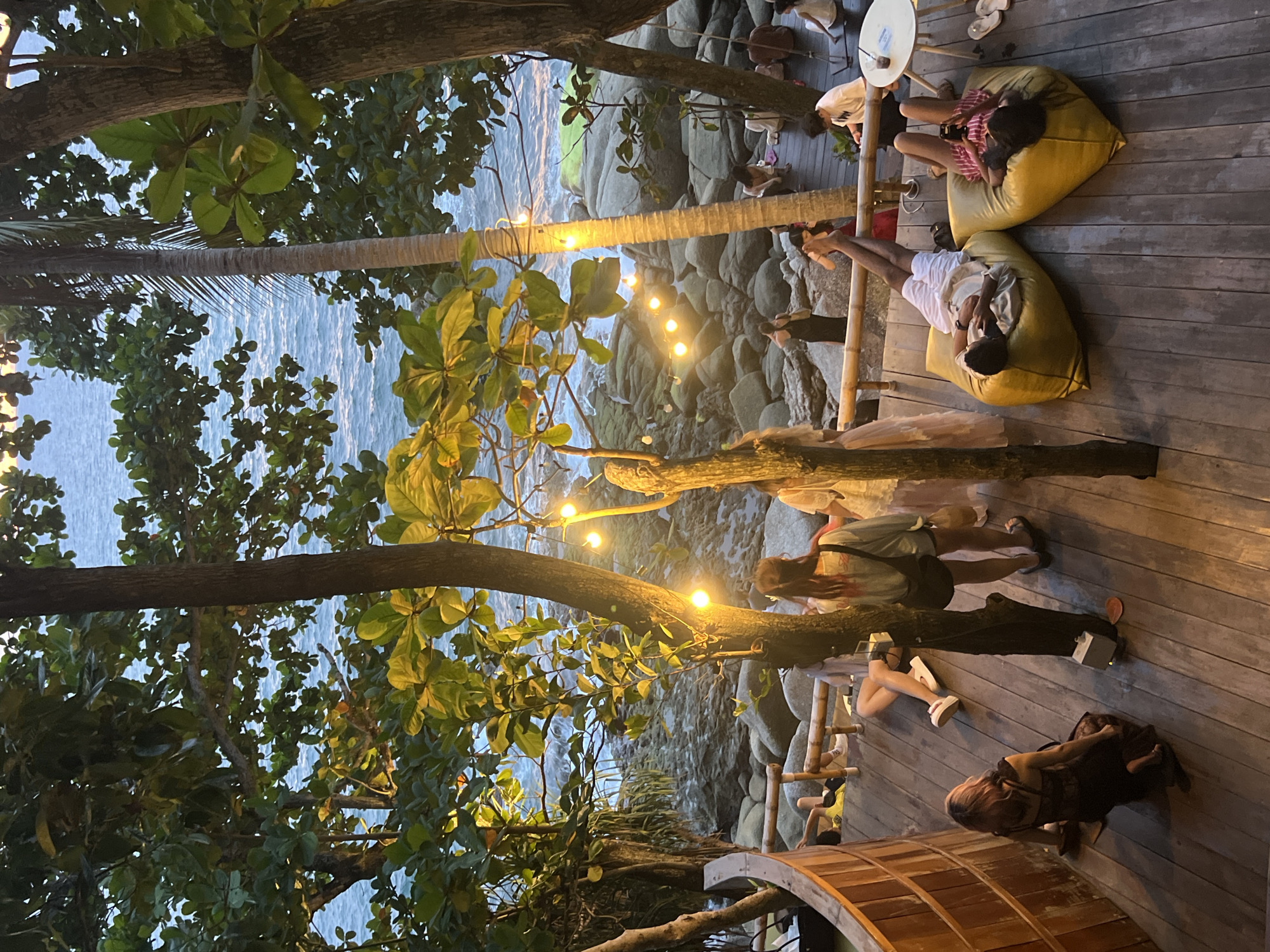 This image captures an outdoor seating area overlooking the ocean in Karon, Thailand. The setting is a wooden deck surrounded by lush trees and a bamboo railing. String lights are strung between trees, illuminating the area. Several people are scattered across the deck. In the foreground, a person with pink hair is kneeling on the wooden planks, facing away from the camera and toward the left. To their right, two people are standing and talking. Further back, two individuals are lounging on large yellow beanbag chairs, one person looking at their phone. In the background, the ocean waves crash against a rocky shoreline. The overall atmosphere suggests a relaxed, casual dining or lounging experience with a scenic ocean view.