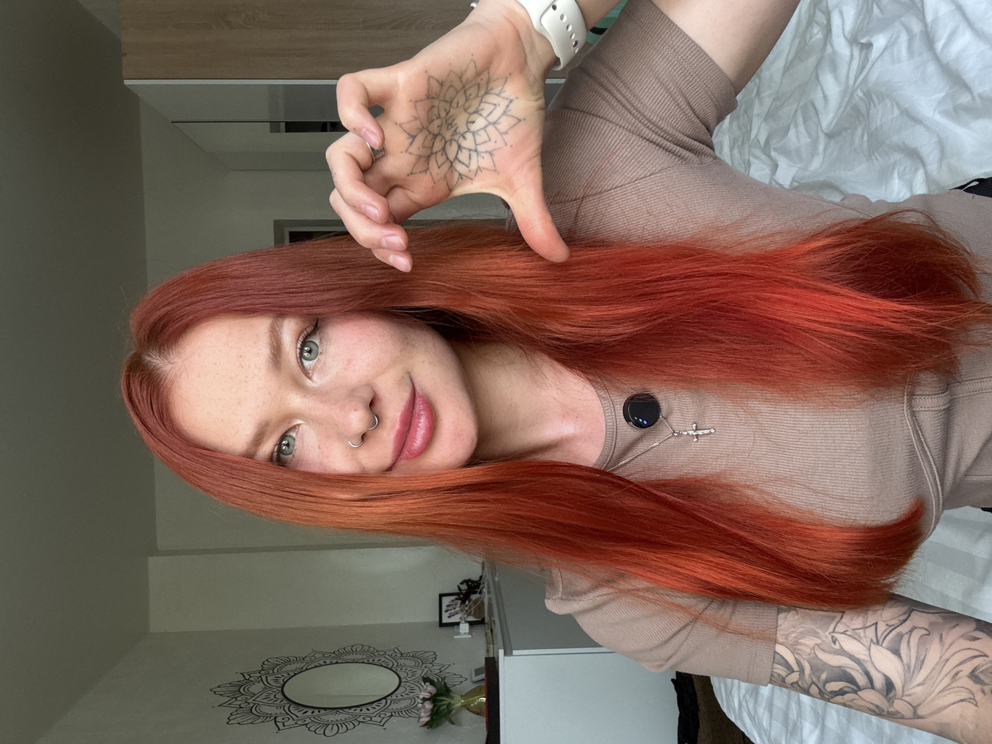 A young woman with red hair and a nose piercing is posing for a selfie in what appears to be a bedroom. She is wearing a beige long-sleeved shirt and a white smartwatch. Her left hand, adorned with a mandala tattoo, is raised and partially obscuring her face. She is also wearing a necklace with a black circular pendant and a small cross. A mirror with a mandala design is visible on the wall in the background, along with a bed with white sheets. The location in the city of Rostov-on-Don, Russia is not discernible from the image.