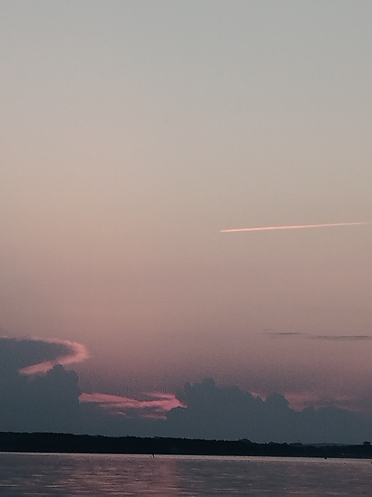 An image taken from the shore of the Kama River in Chaykovsky, Russia, depicts a serene sunset over the water. The sky is a soft gradient of pale pink and lavender, with a faint contrail from an aircraft streaking horizontally across the upper portion of the frame. The lower part of the sky is dominated by silhouetted clouds tinged with pink, suggesting the last rays of the sun. A dark, indistinct shoreline stretches across the bottom of the image, with the calm river reflecting the muted colors of the sky. The overall impression is one of tranquility and natural beauty.