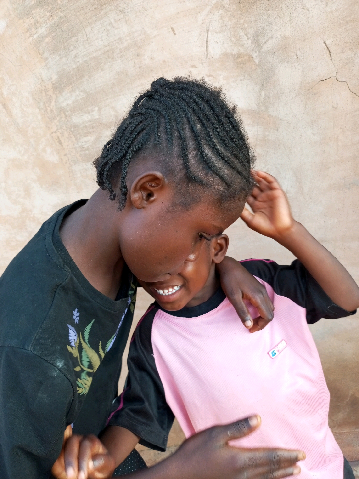Two young Black girls are embracing against a textured, light brown wall. The girl on the left is wearing a black t-shirt with a floral design in shades of purple, green, and white. Her dark hair is braided in cornrows. She is leaning her head down, her face mostly obscured. The girl on the right is wearing a pink t-shirt with black sleeves and a small embroidered logo on the left side of her chest. She is smiling, with her teeth showing, and her right arm is around the other girl's neck. Her left hand is raised near her head. The wall behind them has a rough, uneven surface with some vertical lines and cracks. This image was taken in Tempure, Nigeria.