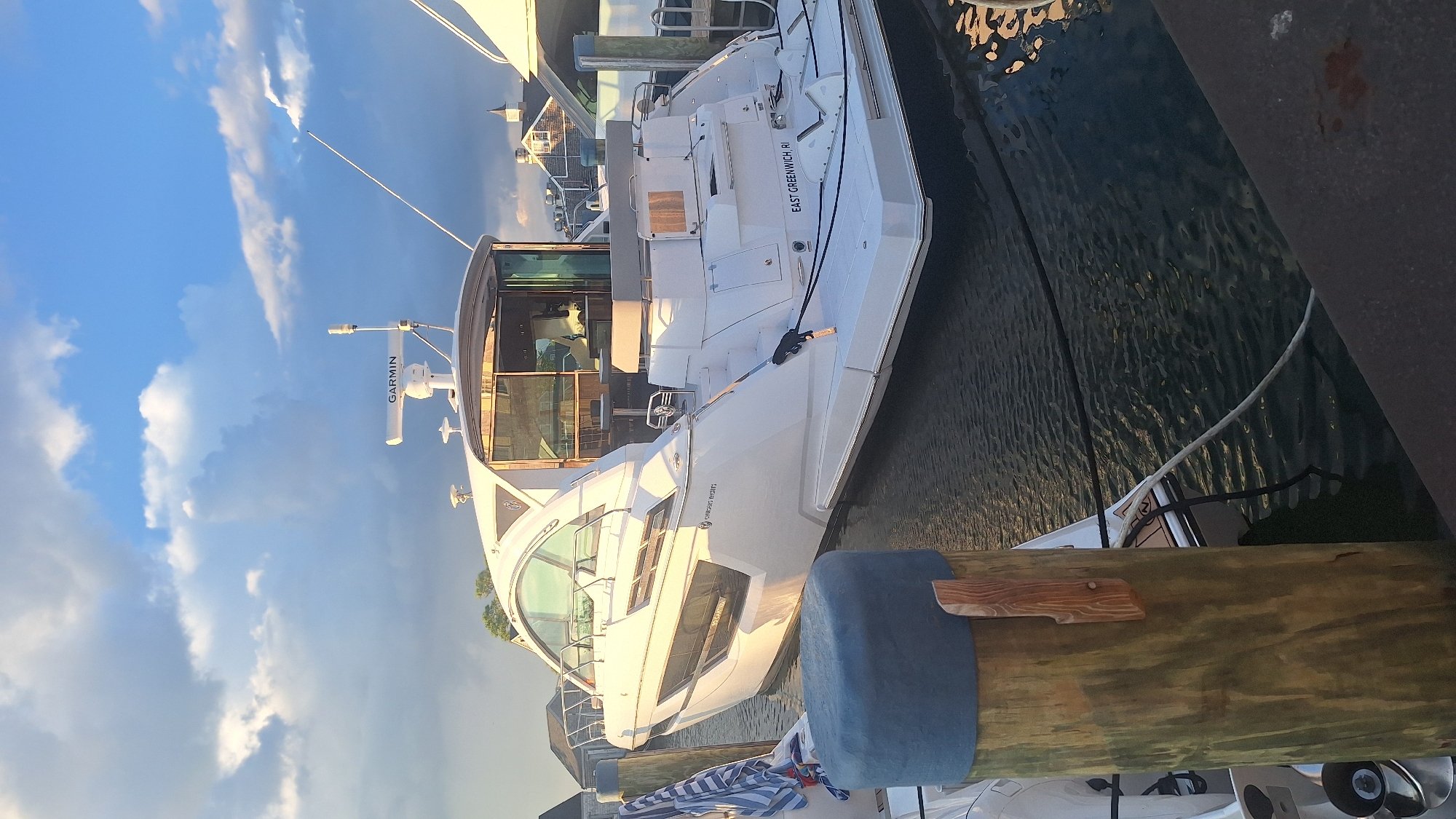 The image captures a white motor yacht docked in a harbor, likely in Nantucket, United States of America, as suggested by the text "EAST GREENWICH, RI" visible on the stern of the boat, indicating a possible registration or homeport. The yacht, featuring a sleek, modern design with large windows, is positioned alongside a wooden dock. In the foreground, a weathered wooden piling with a blue cylindrical fender is prominent. The water is calm, reflecting the sky and the yacht. The sky is partly cloudy with dramatic lighting, suggesting either sunrise or sunset. There are no people clearly visible in the image. Other boats and harbor structures are faintly visible in the background.