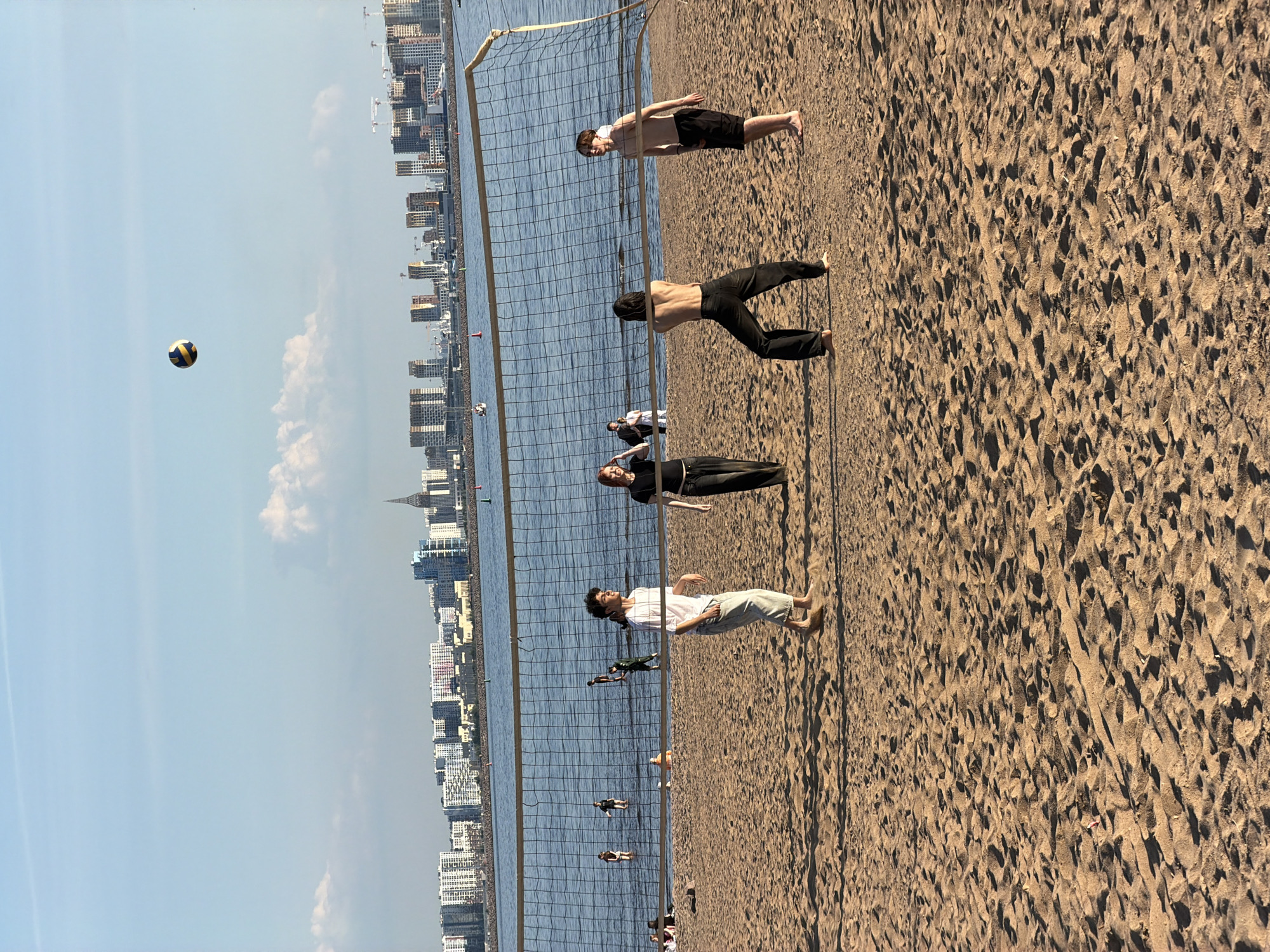 The image depicts a vibrant beach scene in Saint Petersburg, Russia, under clear blue skies. The foreground and midground feature expansive, light-brown sand with visible textures and shadows, indicating bright sunlight. A volleyball net stretches across the scene, planted firmly in the sand and extending into a calm body of water, likely the Gulf of Finland.
Several individuals are present, suggesting a recreational event. On the sand near the net, four people are prominently visible. One person wears a white t-shirt and light pants, another is dressed in a black top and black pants, while two others are bare-chested, wearing dark shorts or pants. All appear barefoot, engaging in beach activities. Further out in the water, numerous smaller figures are visible, some seemingly interacting with the volleyball net, suggesting a game or casual play.
A blue and yellow volleyball is caught mid-air against the expansive sky to the left of the net, reinforcing the theme of a volleyball game. The background is dominated by a dense cityscape, featuring numerous buildings of varying heights, including modern structures and some with visible construction cranes. A distinctive spire-topped building can be seen on the far-right horizon, adding a notable architectural landmark. The water is calm and reflects the bright sky. The overall atmosphere is one of leisure and outdoor activity on a sunny day, with the urban skyline providing a dramatic backdrop to the natural beach environment. No visible text is present in the image.
