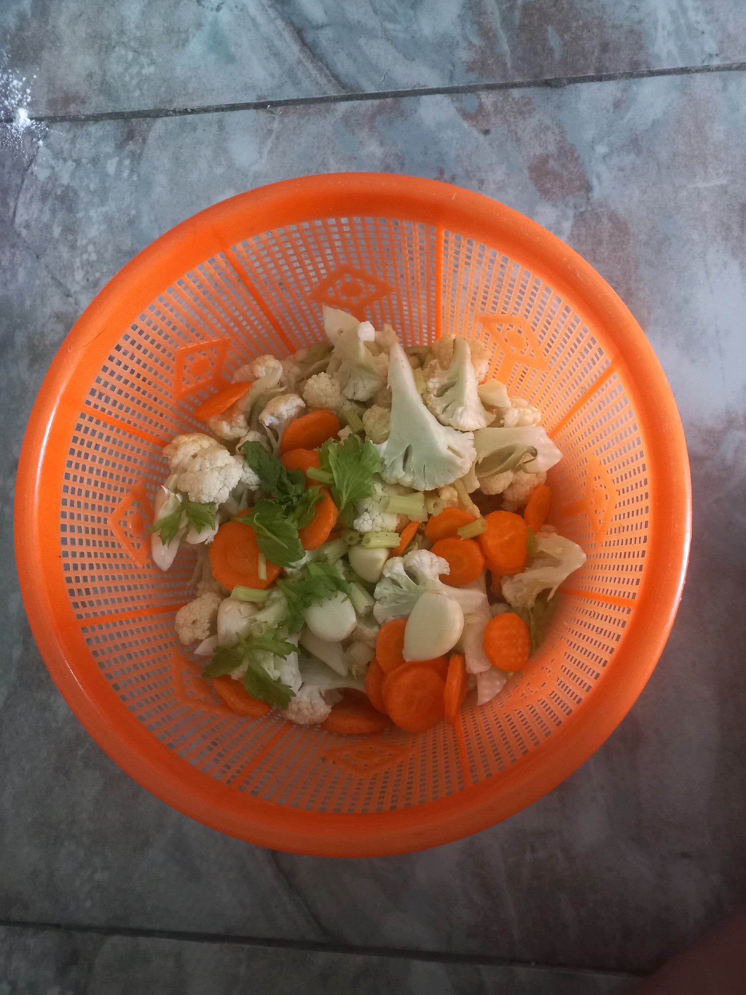 An overhead, medium shot captures a bright orange plastic basket filled with chopped vegetables, including cauliflower florets, sliced carrots, and pieces of celery, resting on a gray tiled floor in Depok, Indonesia. A small pile of white powder is visible on the floor to the left of the basket. The lighting is natural, casting soft shadows and highlighting the texture of the vegetables and the perforations of the basket.