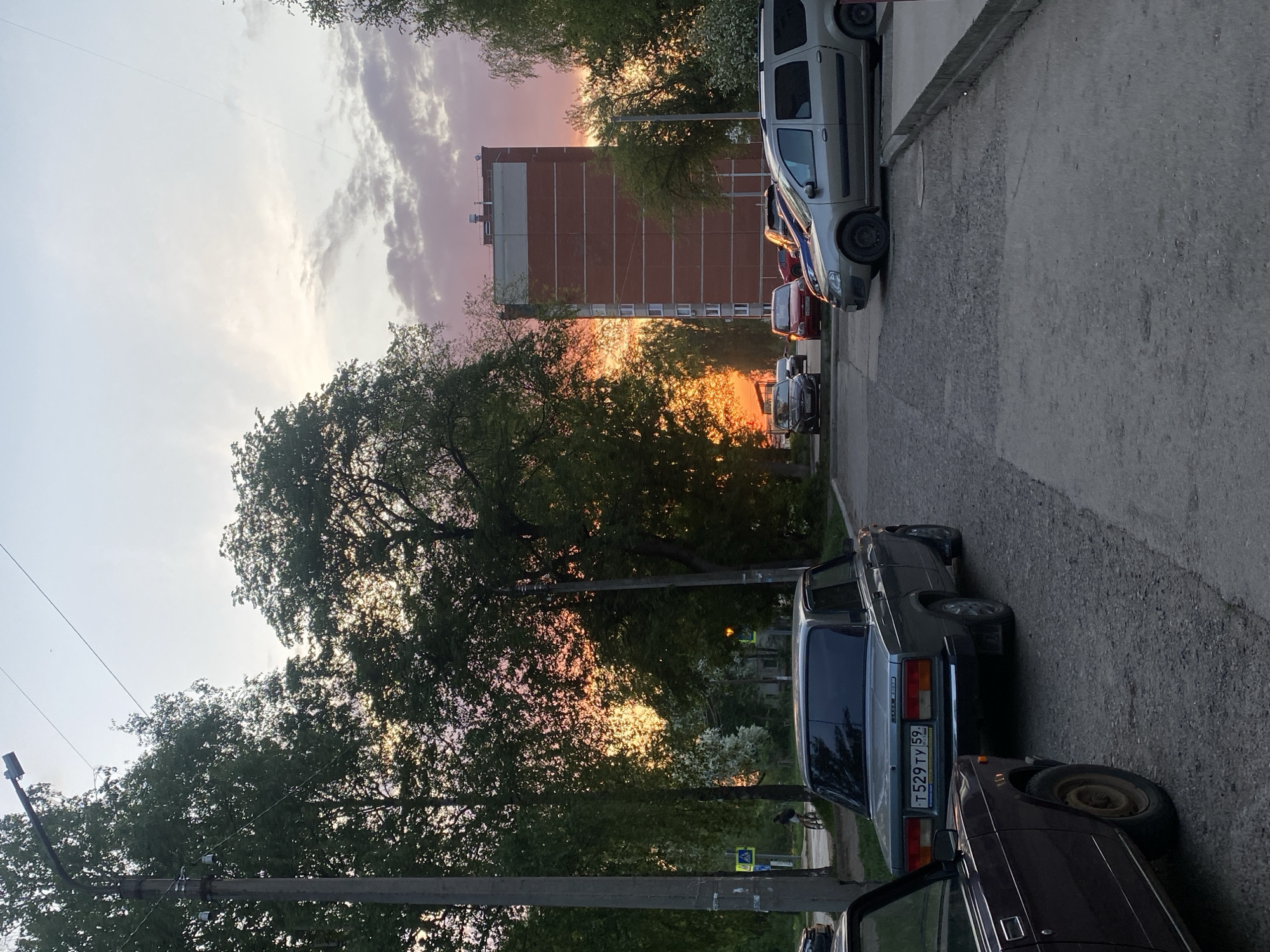 The image depicts a street scene at sunset. In the foreground, two cars are parked next to each other. The license plate on the lighter-colored car reads "T 529 TY 59." In the background, there is a tall brick building and trees. The sun is setting behind the trees, creating an orange glow. Several cars are parked further down the street on the right side of the image. The location is likely in an urban area, but the specific city is unknown. There are no people visible in the immediate foreground.