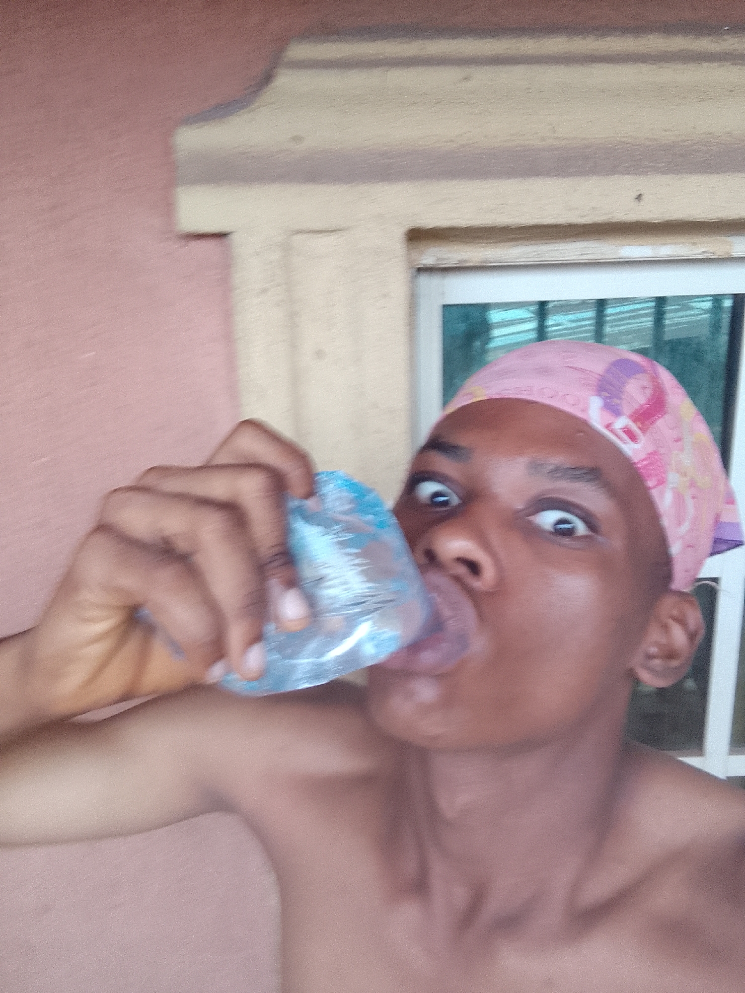A shirtless person wearing a pink headscarf is drinking from a clear plastic bag of liquid. The person is positioned in front of a window with white frames and green glass, with a light pink wall visible on the left. The location is described as Unknown, Unknown.