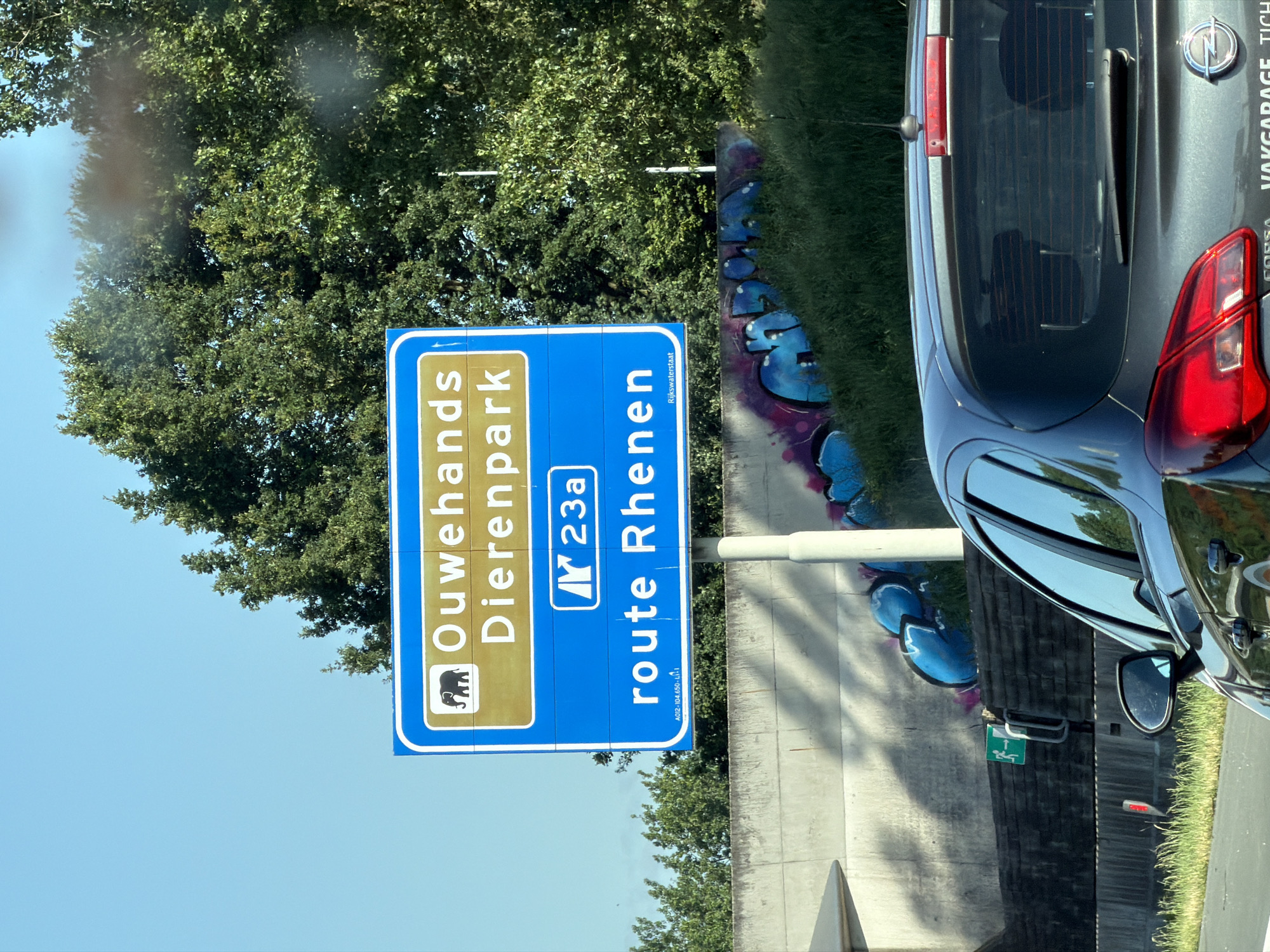 A Dutch road sign for Ouwehands Dierenpark in Rhenen is visible against a bright blue sky and lush green trees. The sign is blue with white text and an elephant graphic. The text reads "Ouwehands Dierenpark" and "route Rhenen." Below the main text, "Rijkswaterstaat" is printed in smaller white letters. The sign is mounted on a white pole. In the foreground, a dark gray car, possibly a hatchback, is driving on a road. Graffiti in blue and purple is visible on a concrete wall behind the sign. To the left of the sign, a green emergency exit sign with a running figure is attached to a metal post. The scene suggests a location in De Klomp, Netherlands, given the reference to Ouwehands Dierenpark.