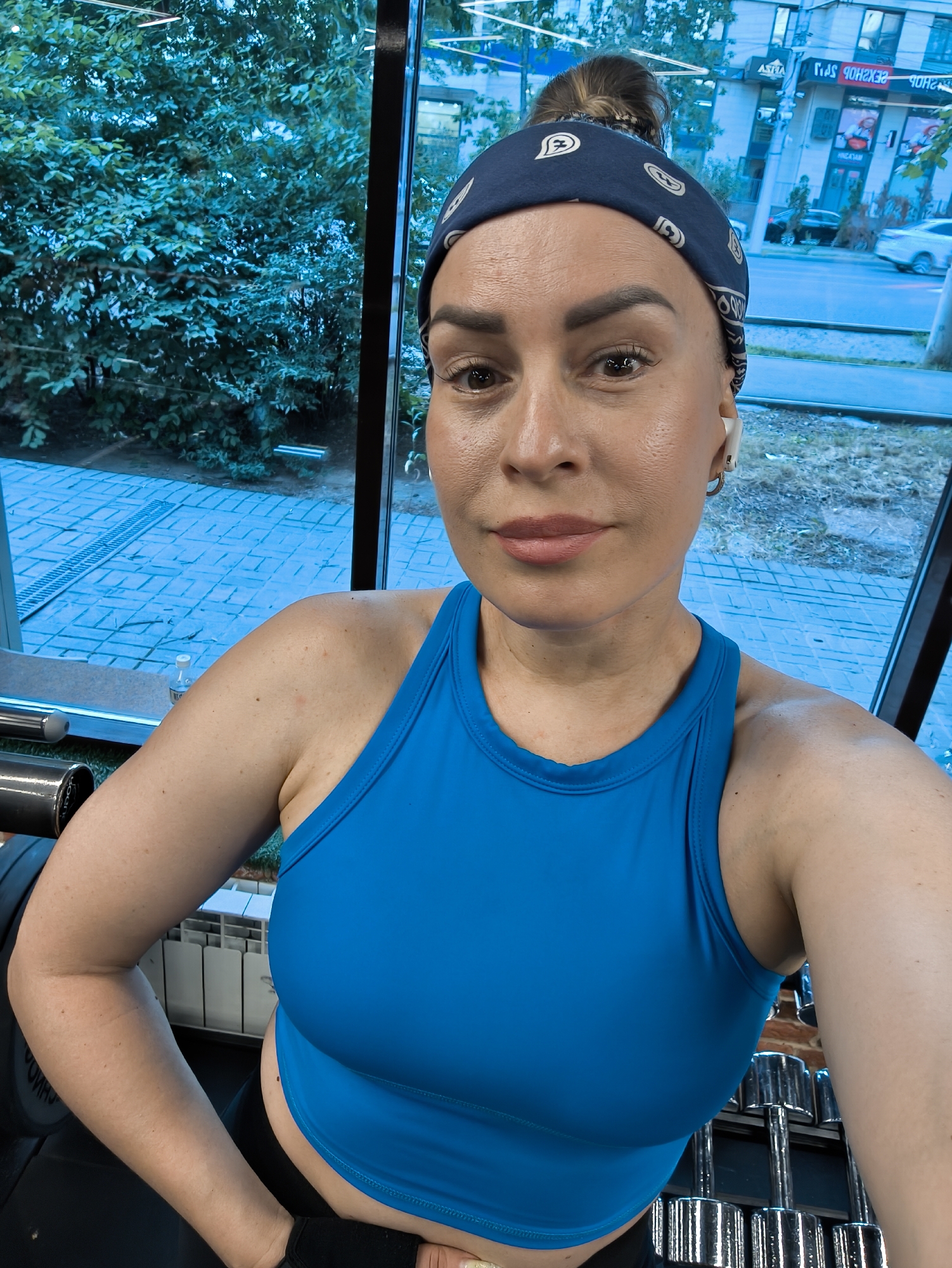 The image is a selfie taken by a woman in a gym, with large windows in the background showing a street scene. The woman is wearing a blue tank top and a bandana. Through the window, buildings and trees are visible. The street appears to be in Almaty, Kazakhstan, as indicated by the Cyrillic text on one of the buildings, which translates to "ASTRA Shopping Center". The woman is looking at the camera, and her expression is neutral. The gym equipment, including dumbbells, is visible in the foreground. The overall lighting suggests it is daytime.