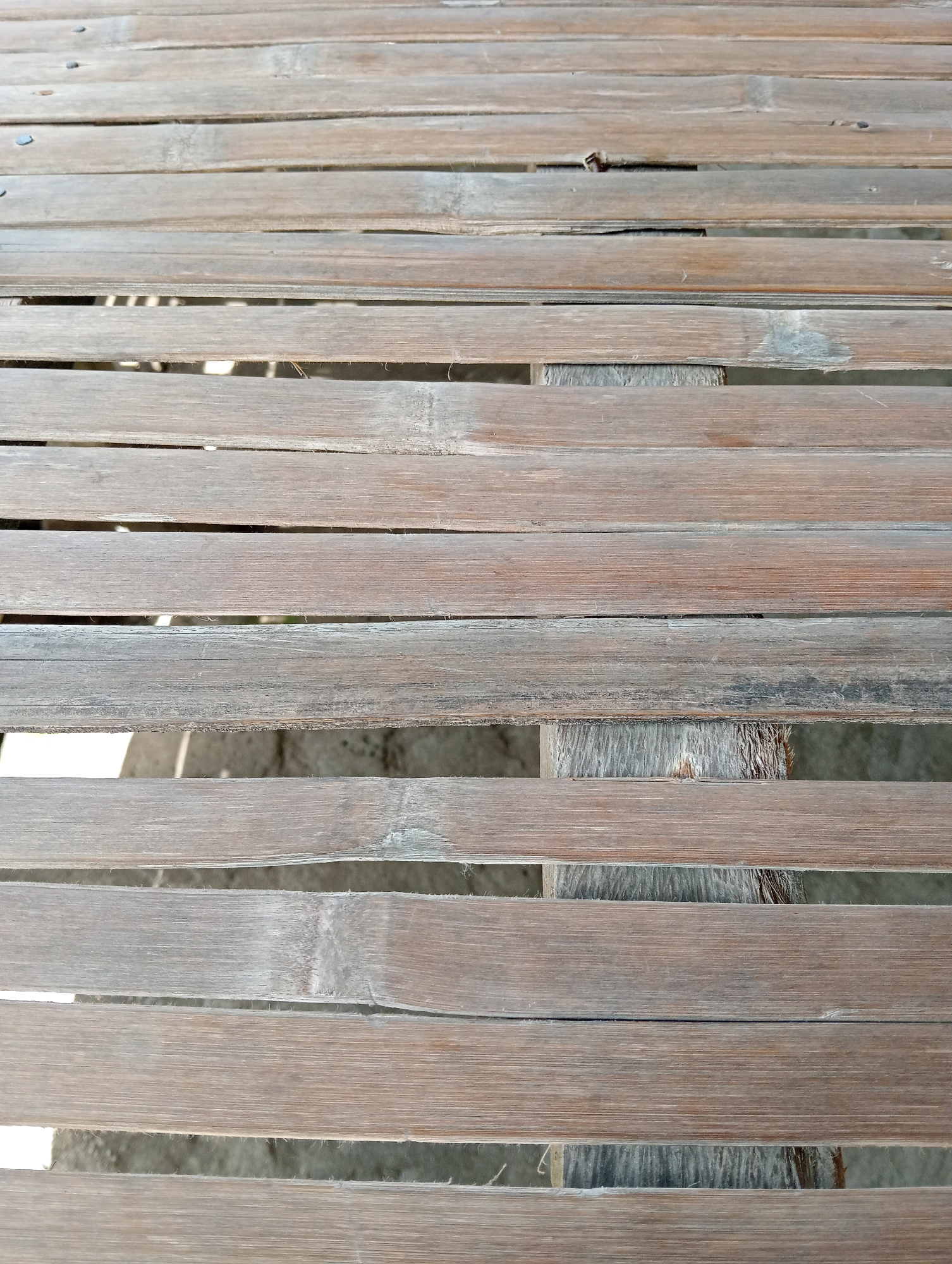 The image shows a close-up view of a weathered wooden surface, likely a bench or table, made of horizontal slats. The wood appears aged and has a textured, dry appearance with visible grain and some lighter, faded areas. There are gaps between the slats, revealing a darker, out-of-focus background that suggests a shaded area or the ground below. The lighting is somewhat uneven, creating subtle shadows and highlights across the surface. The scene is devoid of any people or specific identifying markers that would pinpoint its exact location in Pagerungan Kecil, Indonesia, other than it is an outdoor, potentially rustic setting.
