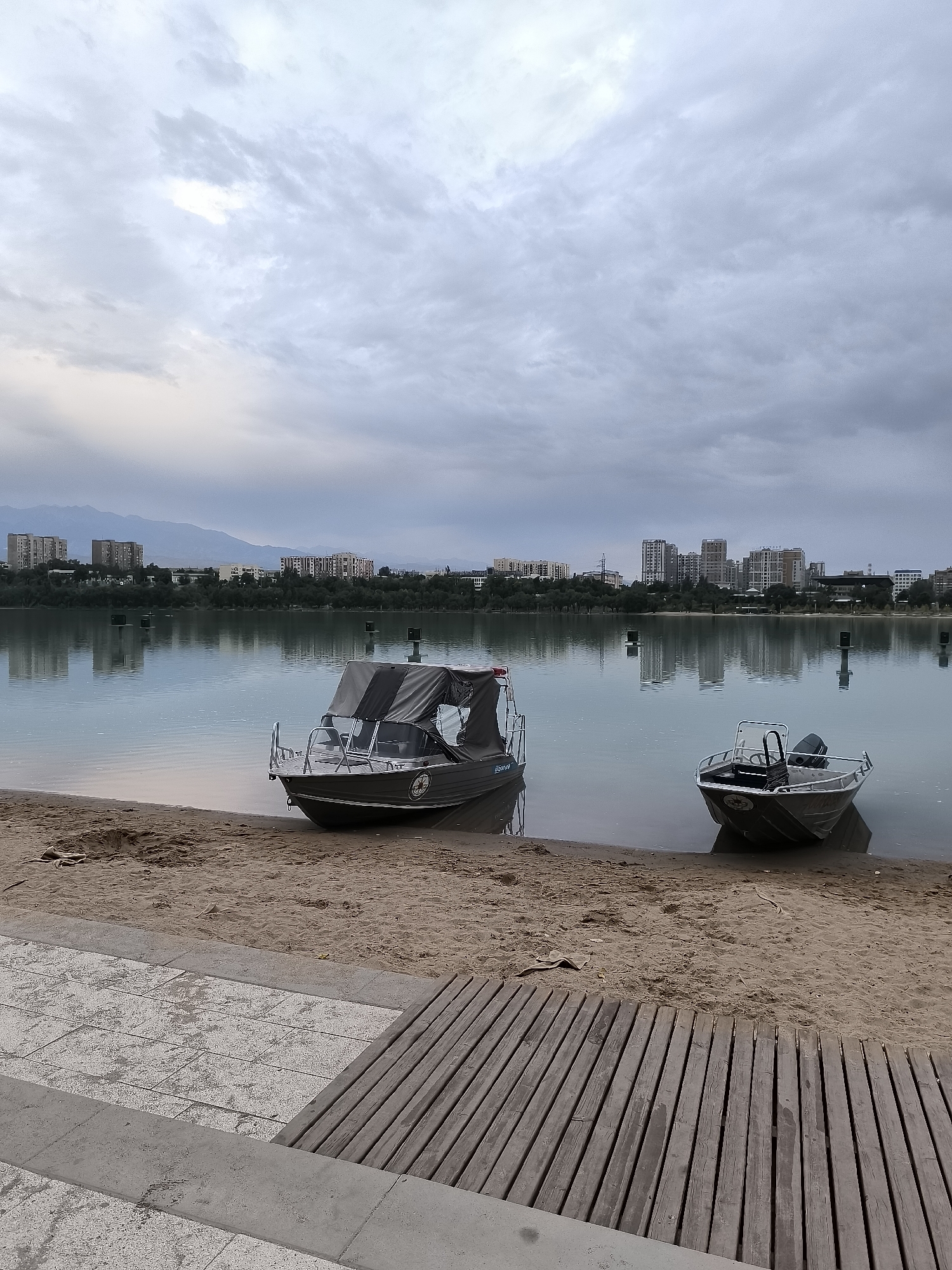 Two small motorboats are pulled up onto a sandy shore in front of a body of water. The boat on the left is larger and has a dark-colored canopy covering most of its interior. The boat on the right is smaller and open. In the background, a city skyline with numerous buildings and a mountain range can be seen across the water under a cloudy sky. The foreground consists of a tiled walkway and a wooden deck. The scene appears to be a park or recreational area.