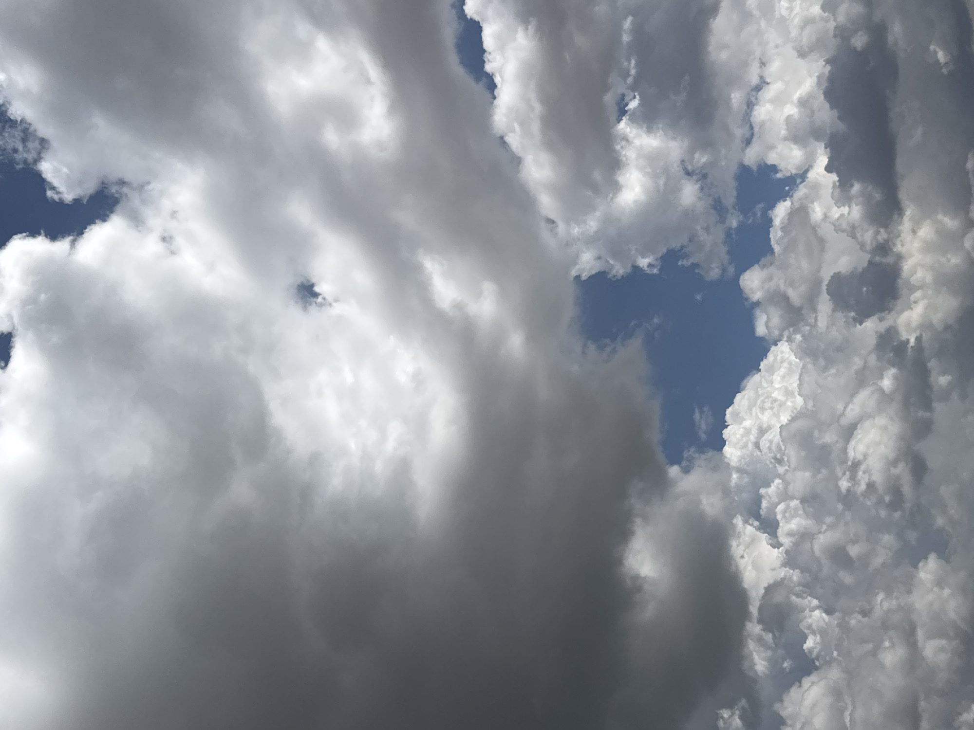 This is a close-up, low-angle shot of a sky filled with dramatic, turbulent clouds. The clouds are predominantly gray and white, with some patches of deep blue sky visible. The lighting suggests it is daytime, with the sun partially obscured by the thick cloud cover, creating a sense of depth and texture. The clouds appear to be moving and forming, with some areas showing bright, illuminated edges, possibly indicating an approaching storm or the aftermath of one. The overall mood is one of grandeur and perhaps a touch of foreboding due to the dark, heavy clouds. The image offers no specific location cues beyond the sky itself, and no text is visible.