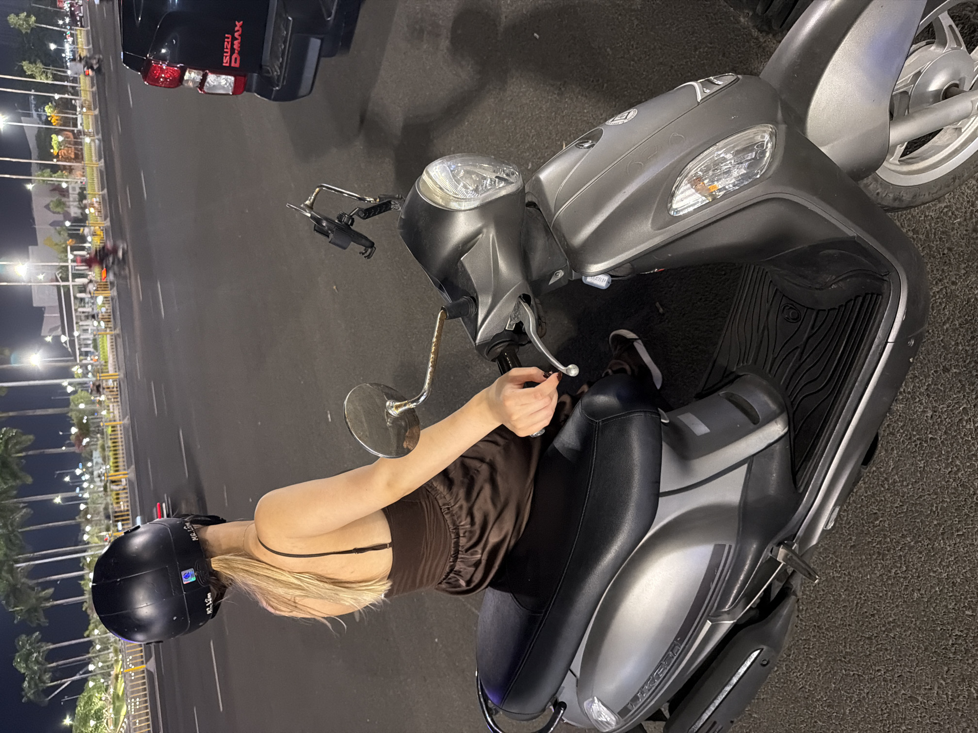 A person wearing a black helmet and a dark brown dress is seated on a grey scooter at night on a city street. The person's back is to the camera, and their blonde hair is visible under the helmet. The scooter has a black seat and chrome handlebars. In the background, palm trees are lit by streetlights, and several vehicles, including a black Isuzu D-MAX truck, are on the road. The scene appears to be in Da Nang, Vietnam, likely near a coastal area given the palm trees.