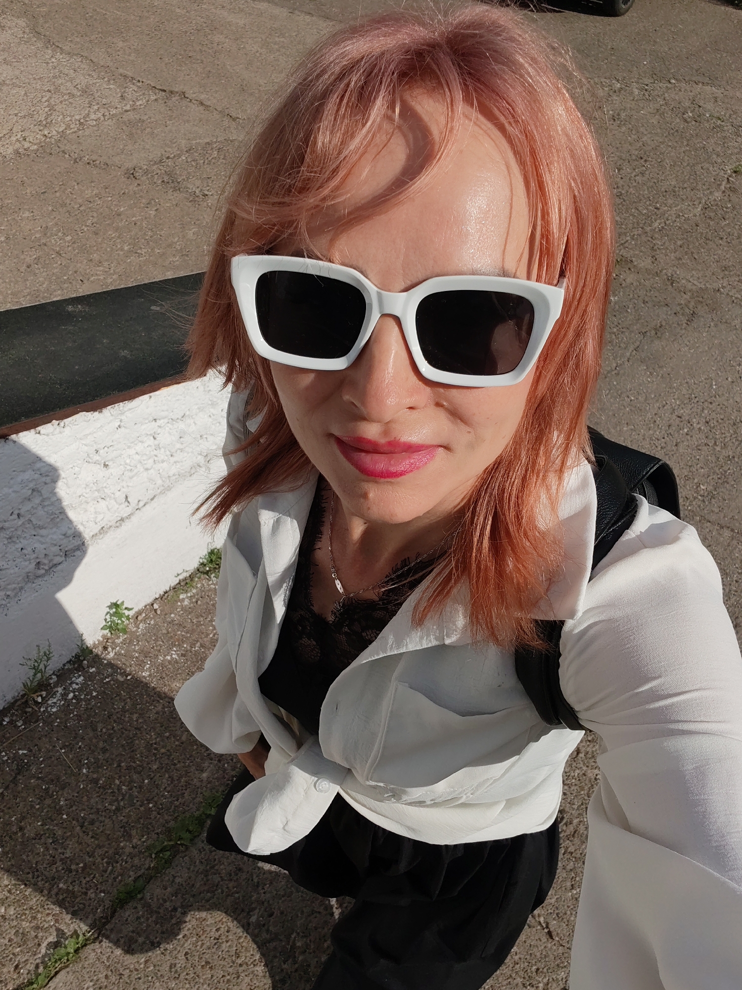 A woman with pinkish-blonde hair and white-framed sunglasses is taking a selfie outdoors. She is wearing a white open shirt over a black lace top, and black pants. A black backpack is visible over her left shoulder. The background consists of a textured concrete surface and a white, rough-textured wall with some greenery at its base. The lighting suggests it is daytime. The overall scene appears to be an outdoor urban or suburban environment, possibly near a building or a park. The location is identified as Chaikovsky, Russia.