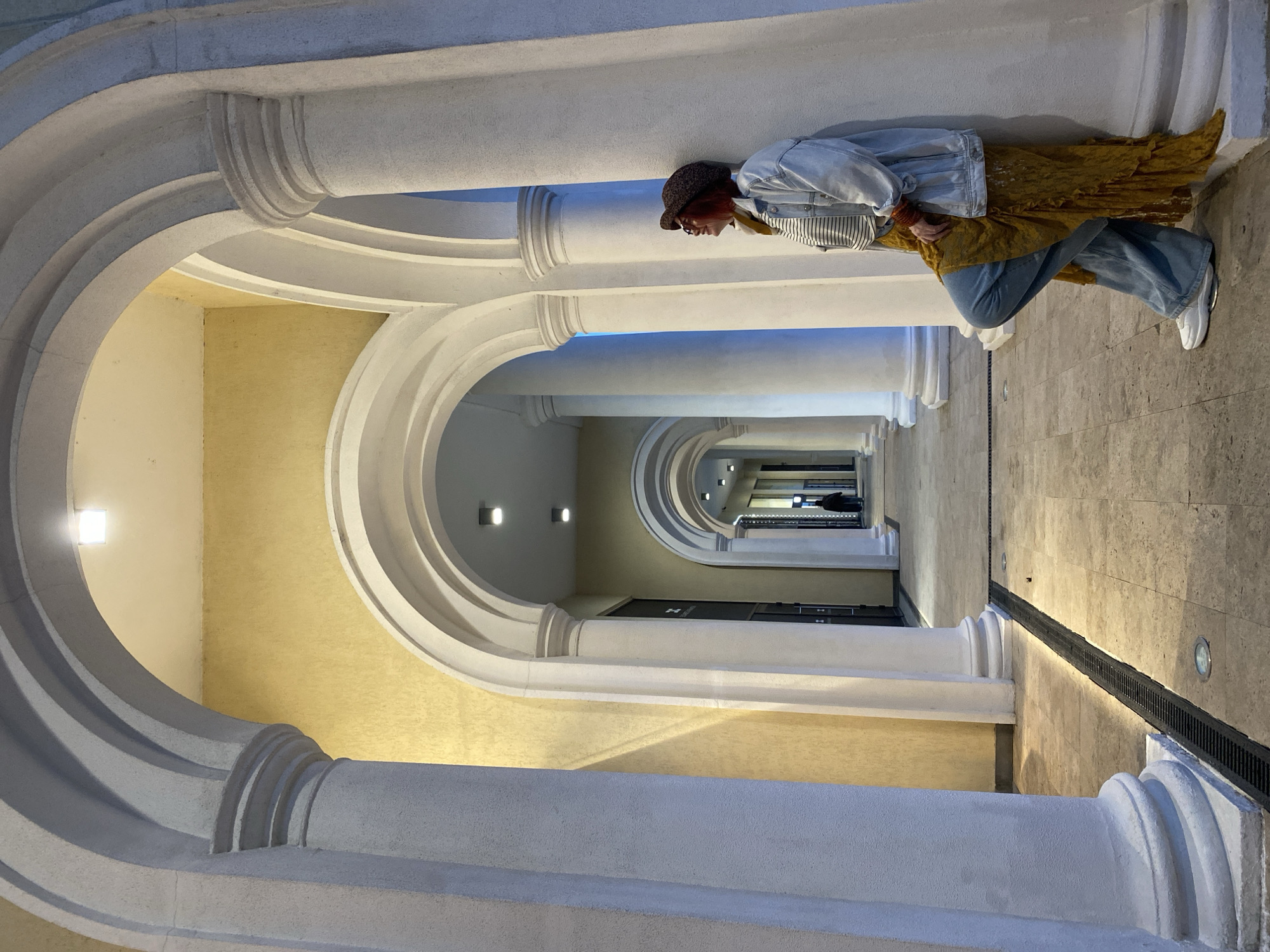 The image shows a hallway with a series of arched columns, leading into the distance, in a building in Sochi, Russia. The walls are painted a light yellow, and the columns are white. The floor is tiled. A person is standing in the hallway, wearing a hat, denim jacket, striped shirt, gold colored skirt, and blue jeans. They are facing away from the camera, towards the end of the hallway.