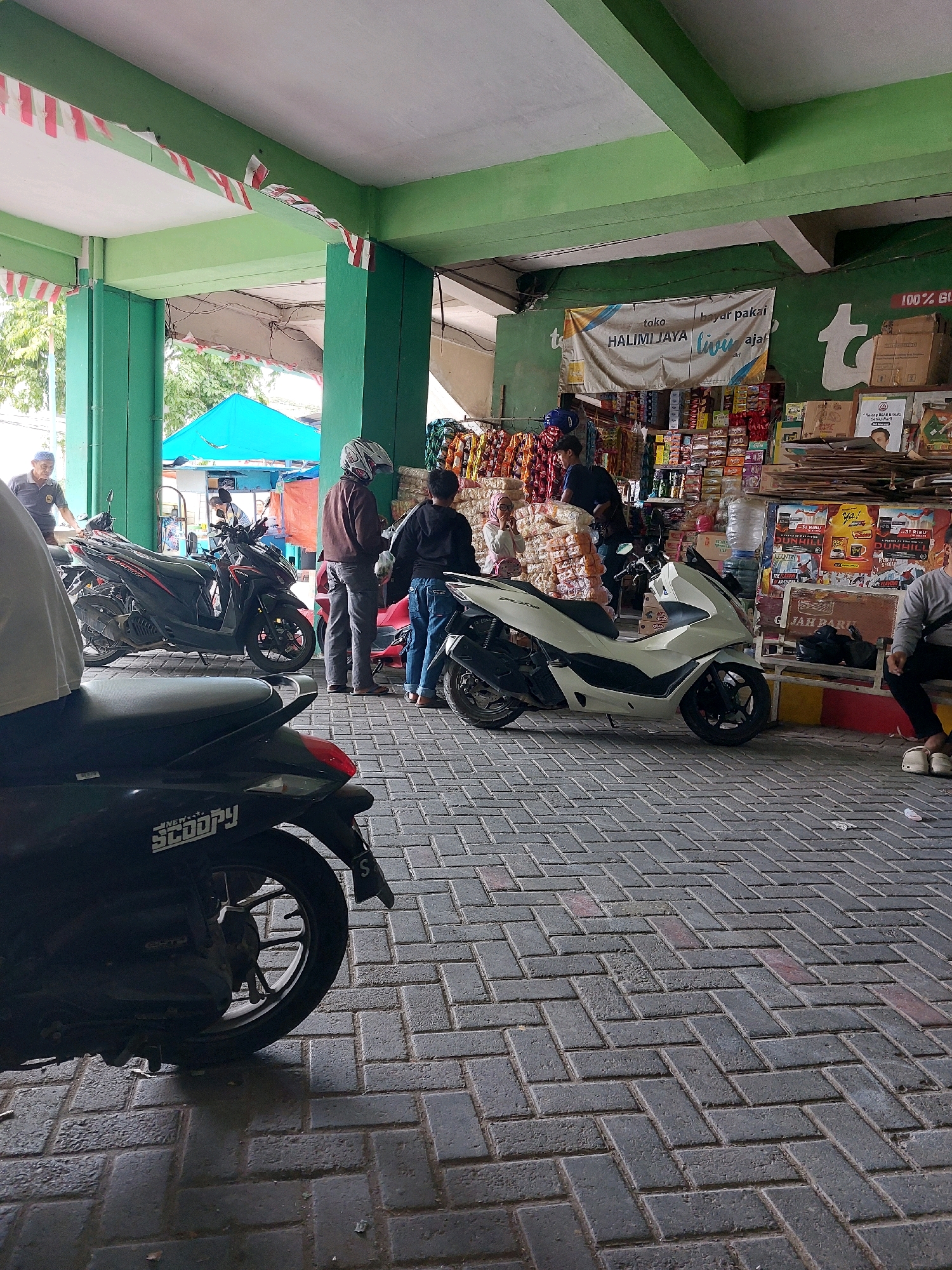 An outdoor scene in Sumuragung, Indonesia, shows a section of a marketplace or convenience store with an overhang supported by green columns. Red and white Indonesian flags are draped along the edge of the overhang. A variety of motorcycles are parked on a brick-paved ground. In the background, shelves are stocked with goods, and several people are present. One person is wearing a helmet and a brown jacket, standing near a white motorcycle. Another person in a black hoodie is next to them. A man in a blue shirt and cap is walking towards the left side of the frame. A woman in a pink headscarf is visible near a stack of bagged goods. A white motorcycle is parked prominently in the foreground. A black motorcycle is partially visible on the far left. A person in a grey long-sleeved shirt is sitting on a bench on the right side of the image. The overall atmosphere suggests a busy commercial area.