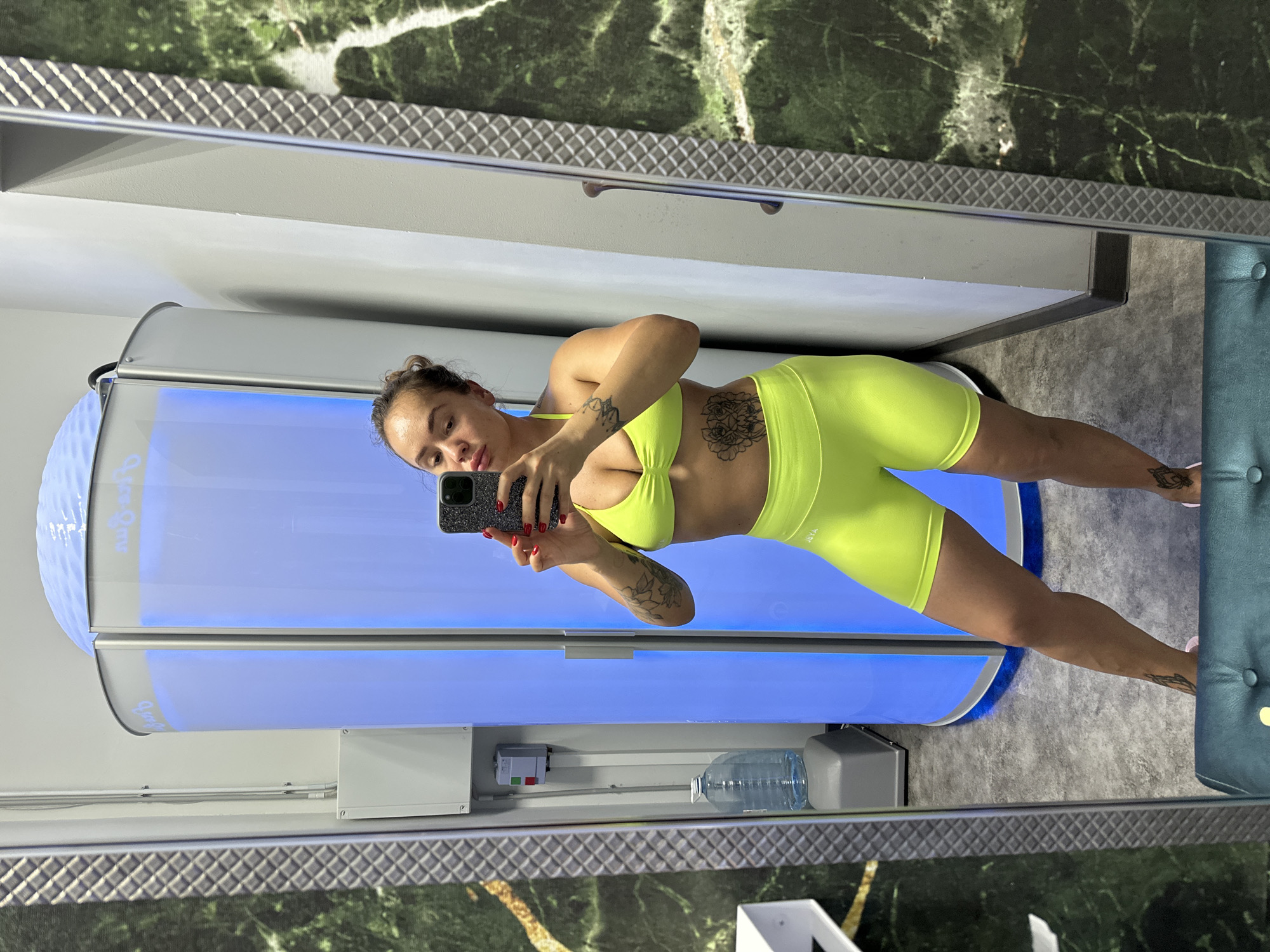 A woman is taking a mirror selfie in what appears to be a tanning salon. She is wearing a neon yellow sports bra and matching shorts. She has tattoos on her arm and torso. In the background, there is a blue tanning booth, a water cooler, and a wall with a marble-like pattern. The floor is gray.