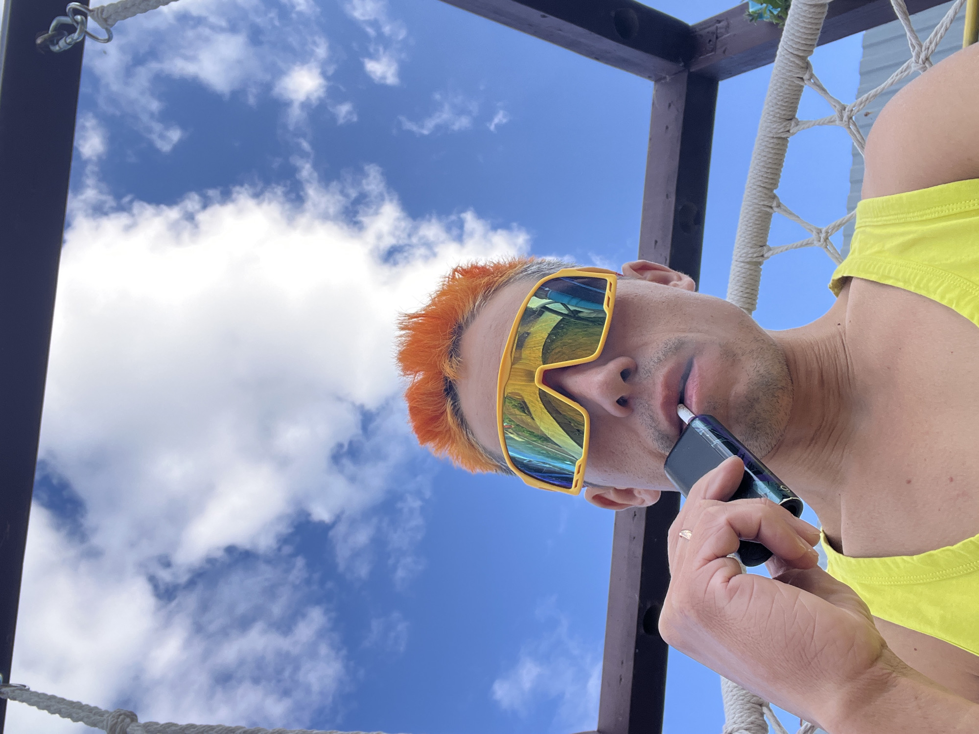 A man with bright orange hair and yellow, mirrored sunglasses is holding a dark electronic smoking device to his mouth. He is wearing a bright yellow tank top and is seated in a rope-strung chair. The background is a clear blue sky with fluffy white clouds. The scene appears to be outdoors, possibly in a recreational area like a park or a backyard, given the presence of the chair and what looks like a wooden frame above. The specific location in Chaikovsky, Russia, is not discernible from the visual information provided in the image.