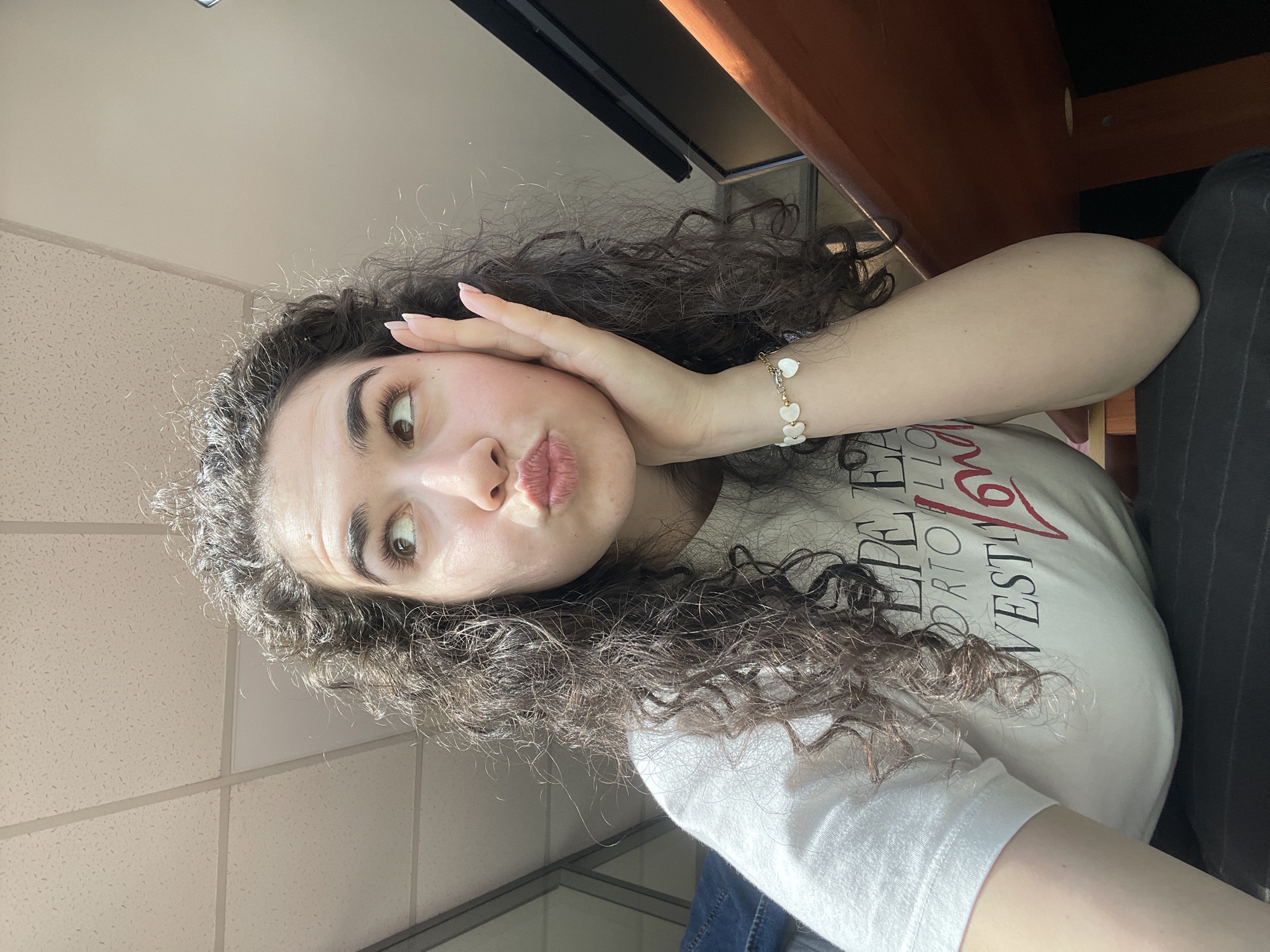 The media file features a young woman with long, dark, curly hair taking a selfie indoors in Sofia, Bulgaria. She is positioned with her head tilted, looking to her left, and her lips are pursed in a playful "kiss face" or pout. Her left hand is raised to her cheek, with fingers extended and light-colored nail polish visible. She wears a delicate bracelet on her left wrist, adorned with multiple small, white, heart-shaped charms.
She is dressed in a light-colored, likely white, t-shirt. Partially visible text on the shirt includes segments like "PE", "ORTO", and "WEST", accompanied by a prominent, stylized red character that resembles a 'V' or '6'.
The background indicates an indoor setting, likely an office or classroom, with a white acoustic ceiling featuring grid lines visible above and to her right. A textured wall is present to her right. To her left, dark wooden furniture, possibly a desk or shelf, along with a dark rectangular object that could be a monitor, are visible. A portion of dark, possibly striped, fabric is also seen on the far right. The bright illumination suggests the photograph was taken during the daytime.
