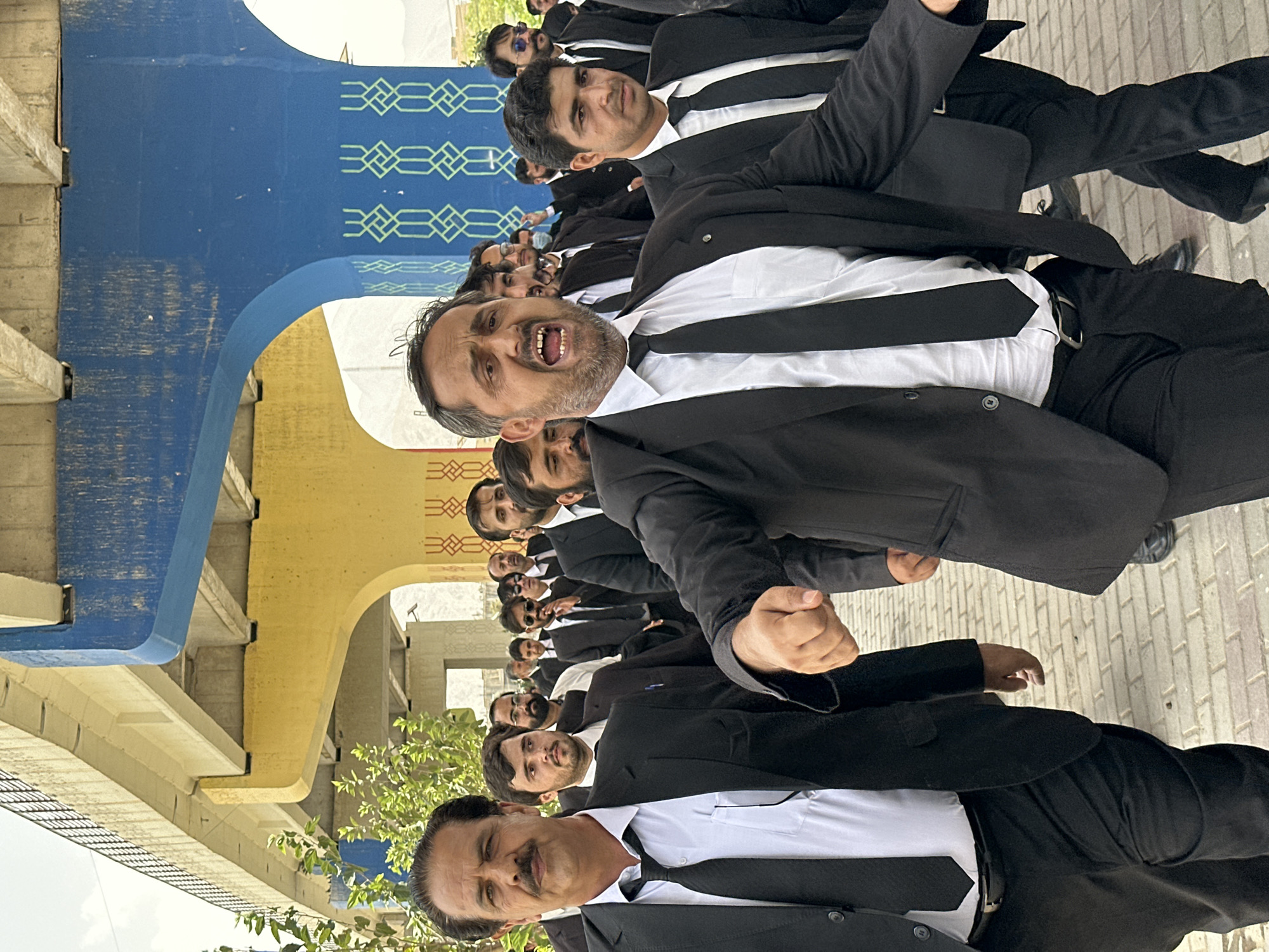 The image shows a group of men in black suits and ties, walking outdoors under an overpass. The overpass has a yellow underside and blue painted pillars with geometric patterns. The ground is paved with light-colored bricks. In the foreground, a man with a mustache is looking forward, with his mouth slightly open. Another man in the foreground is shouting, with his mouth wide open and his right arm raised. Behind them, other men in similar attire are walking. Trees are visible to the left of the frame, and a mountainous landscape can be seen in the background. The overall scene suggests a procession or demonstration.